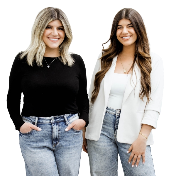 Two women smiling, standing side by side, one in a black top and jeans, the other in a white blazer and jeans.