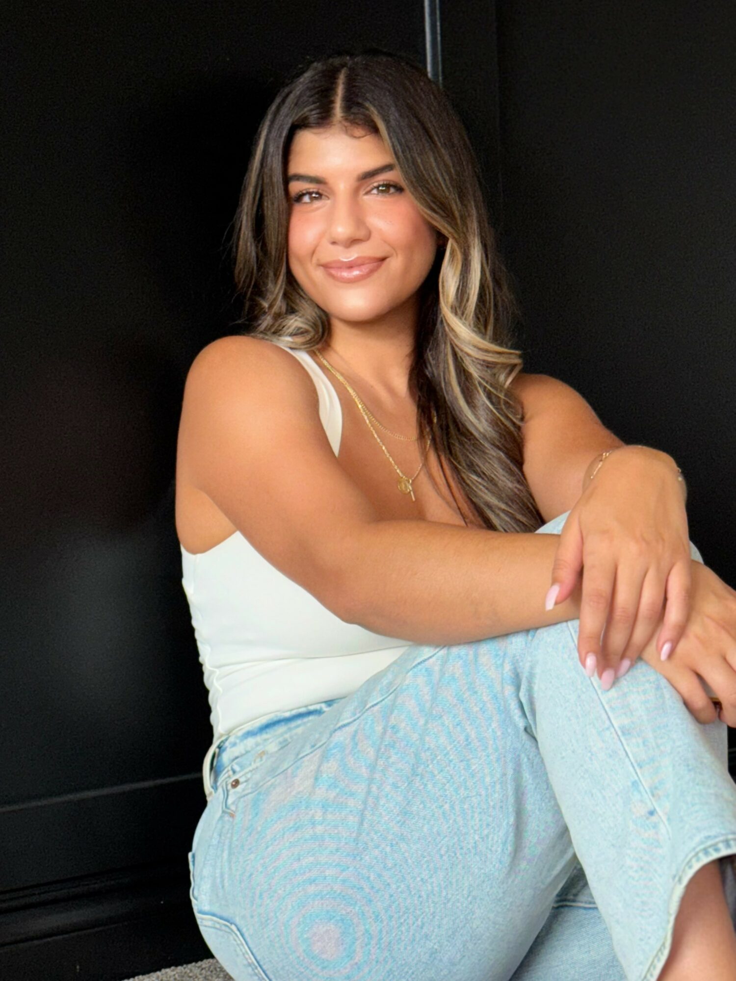 Young woman with long wavy hair, smiling, sitting with arms crossed, wearing a white top and light blue jeans.