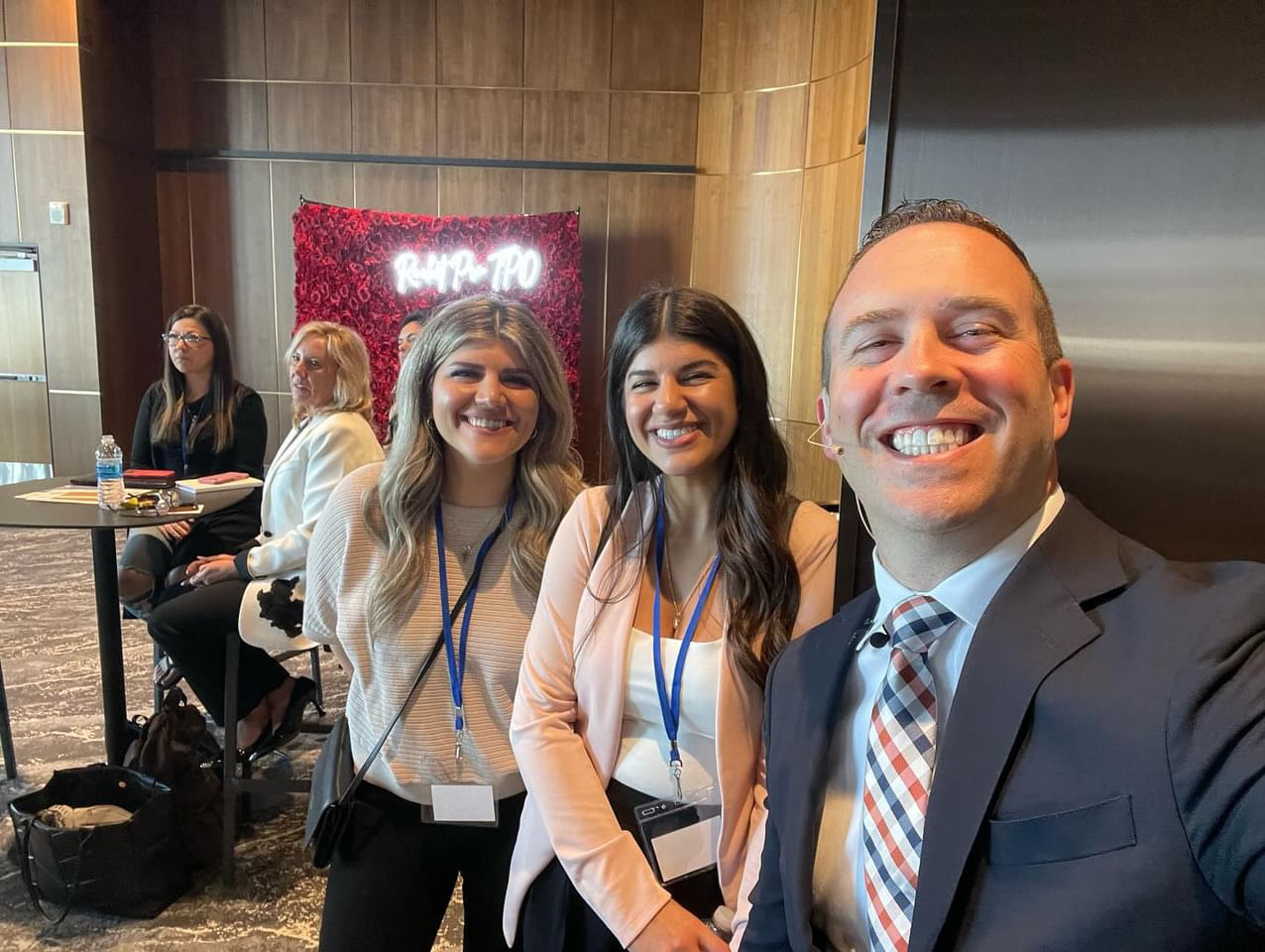 Three people smiling for a selfie in a conference room with a red illuminated sign in the background.