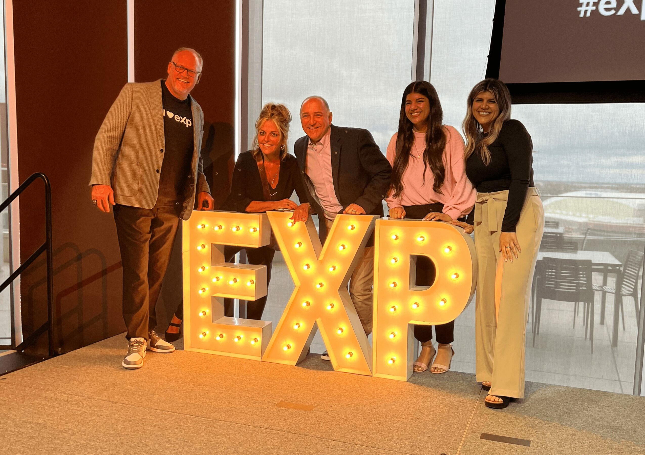 Five people stand behind illuminated letters spelling 'EXP' at an indoor event, with a window and screen in the background.