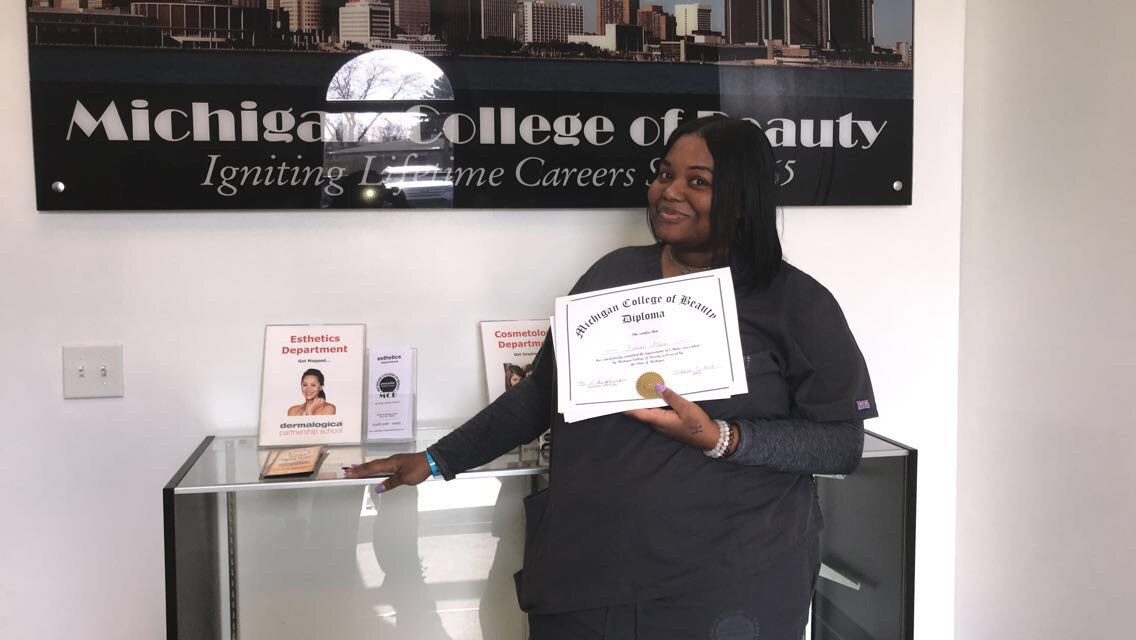 Woman holding a diploma in front of a Michigan College of Beauty sign, smiling, standing next to a glass display case.