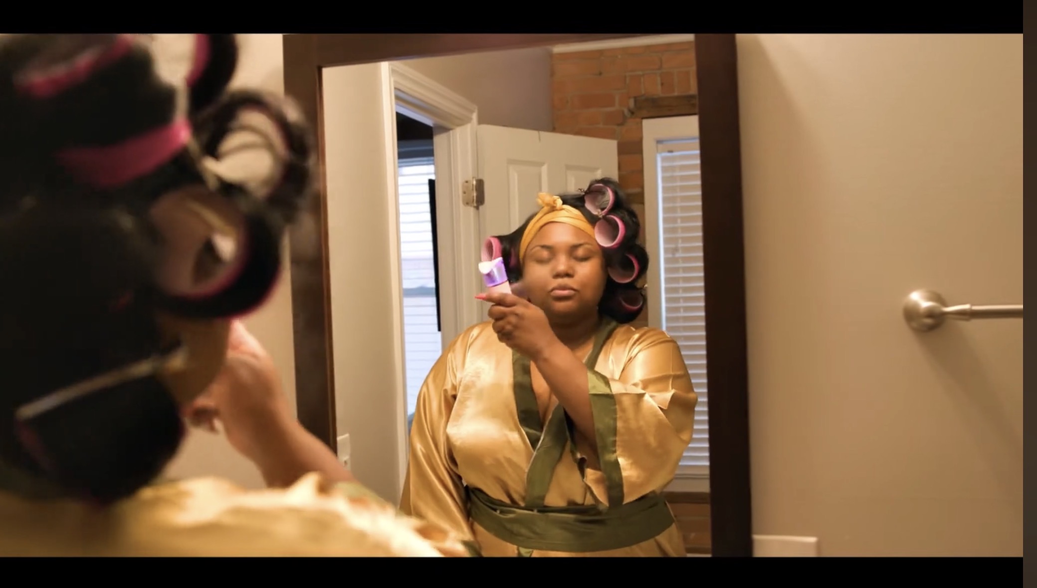 Woman with hair rollers looking at mirror, holding a phone, in a room with a door and mirror.