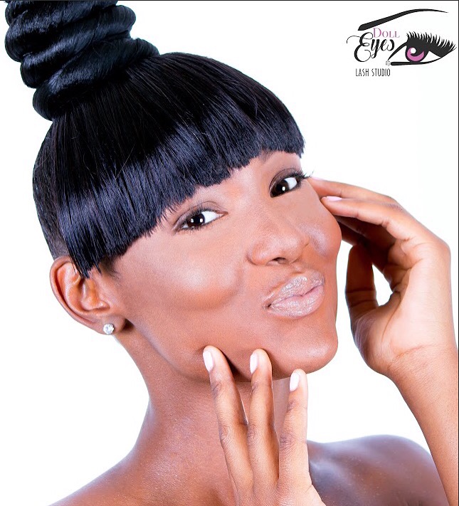 Woman with styled hair, makeup, and earrings, touching her face with one hand, smiling slightly, against a white background.