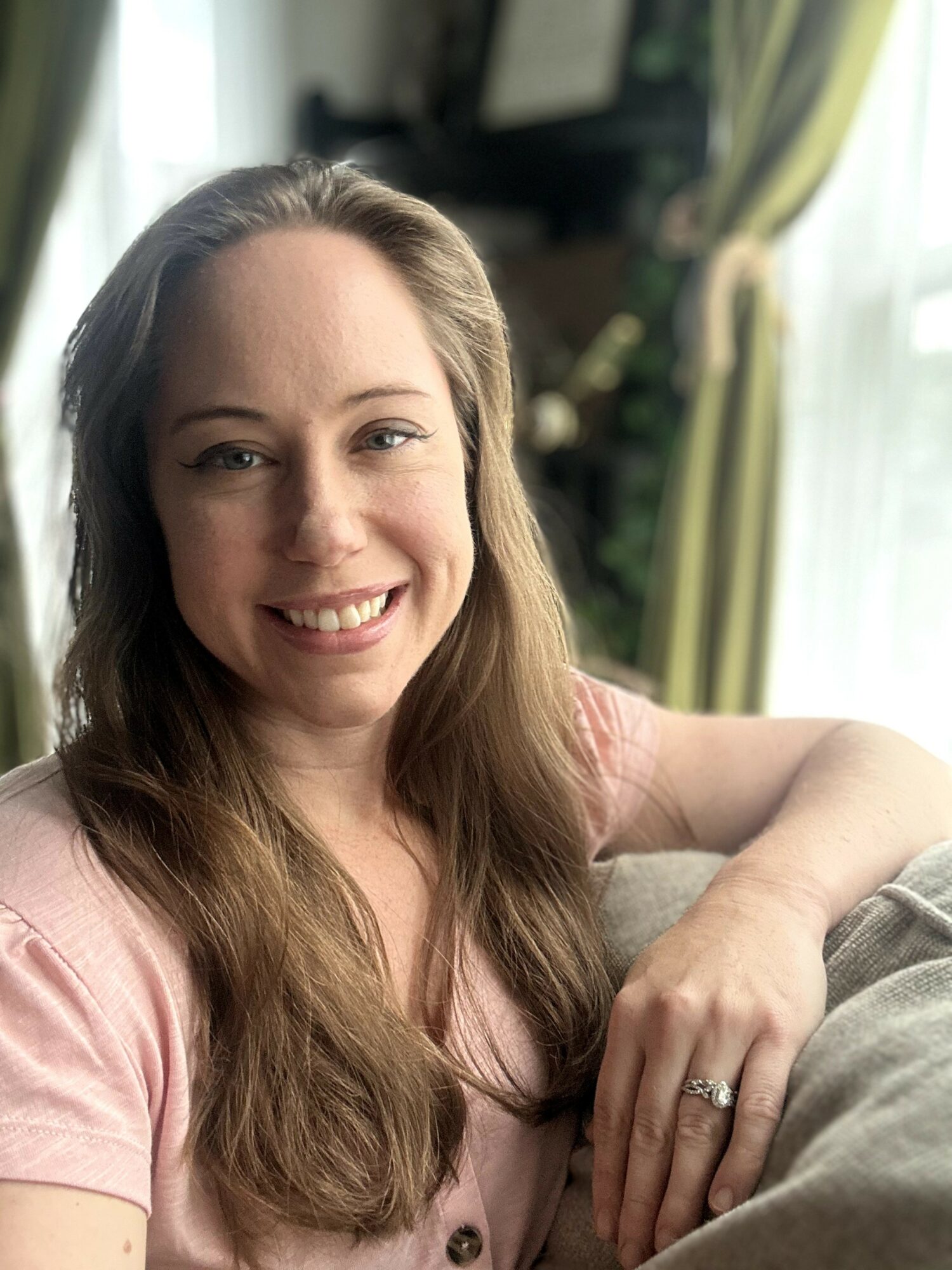Smiling woman with long hair sitting on a couch in a room with green curtains and sunlight.