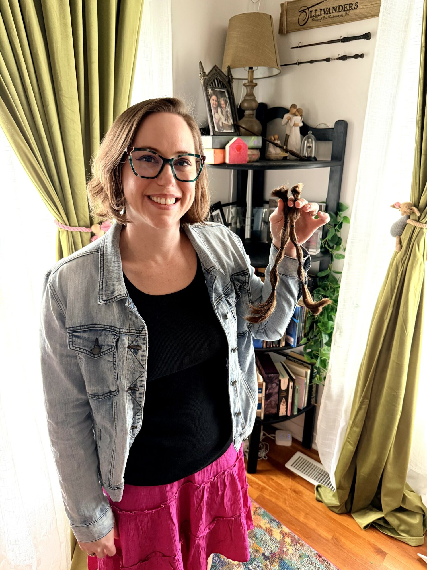 Woman with glasses smiling, holding a camera, standing in a room with green curtains and a bookshelf.