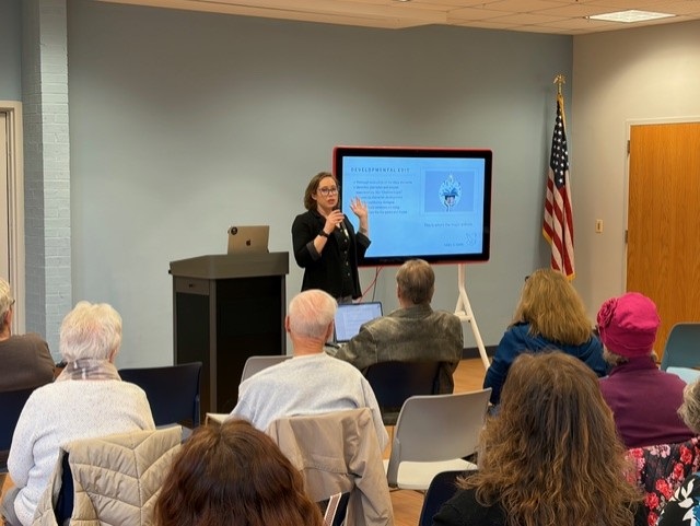 Woman giving a presentation in a room with an audience, a screen displaying a slide, and an American flag, with a podium nearby.