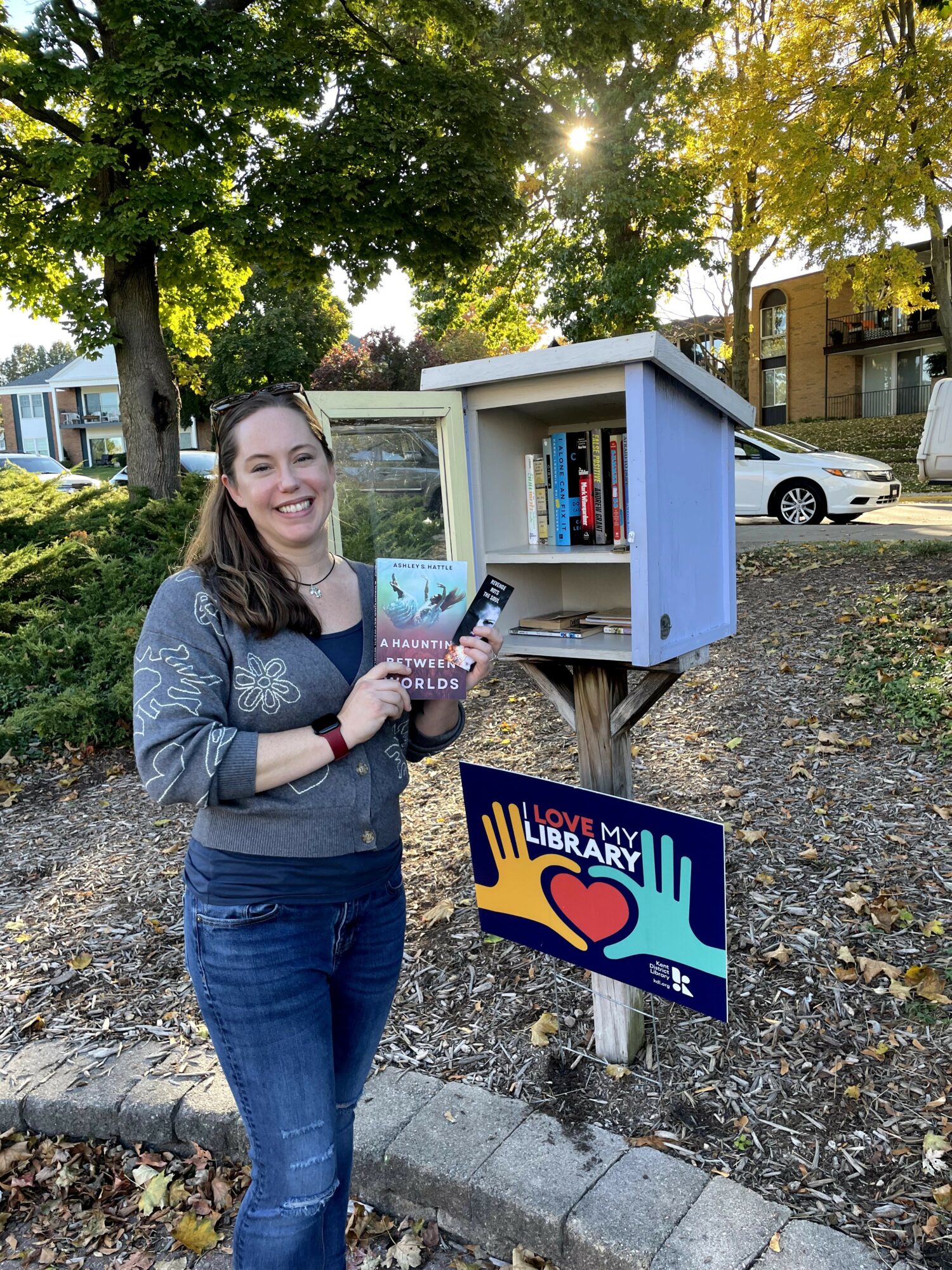 Smiling woman holding a book in front of a small outdoor library box on a sidewalk, with trees and buildings in the background.