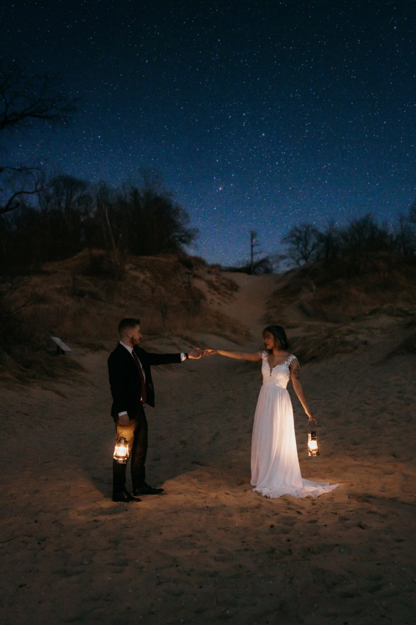 A couple holding lanterns stands on a sandy landscape under a starry night sky, reaching out to each other.