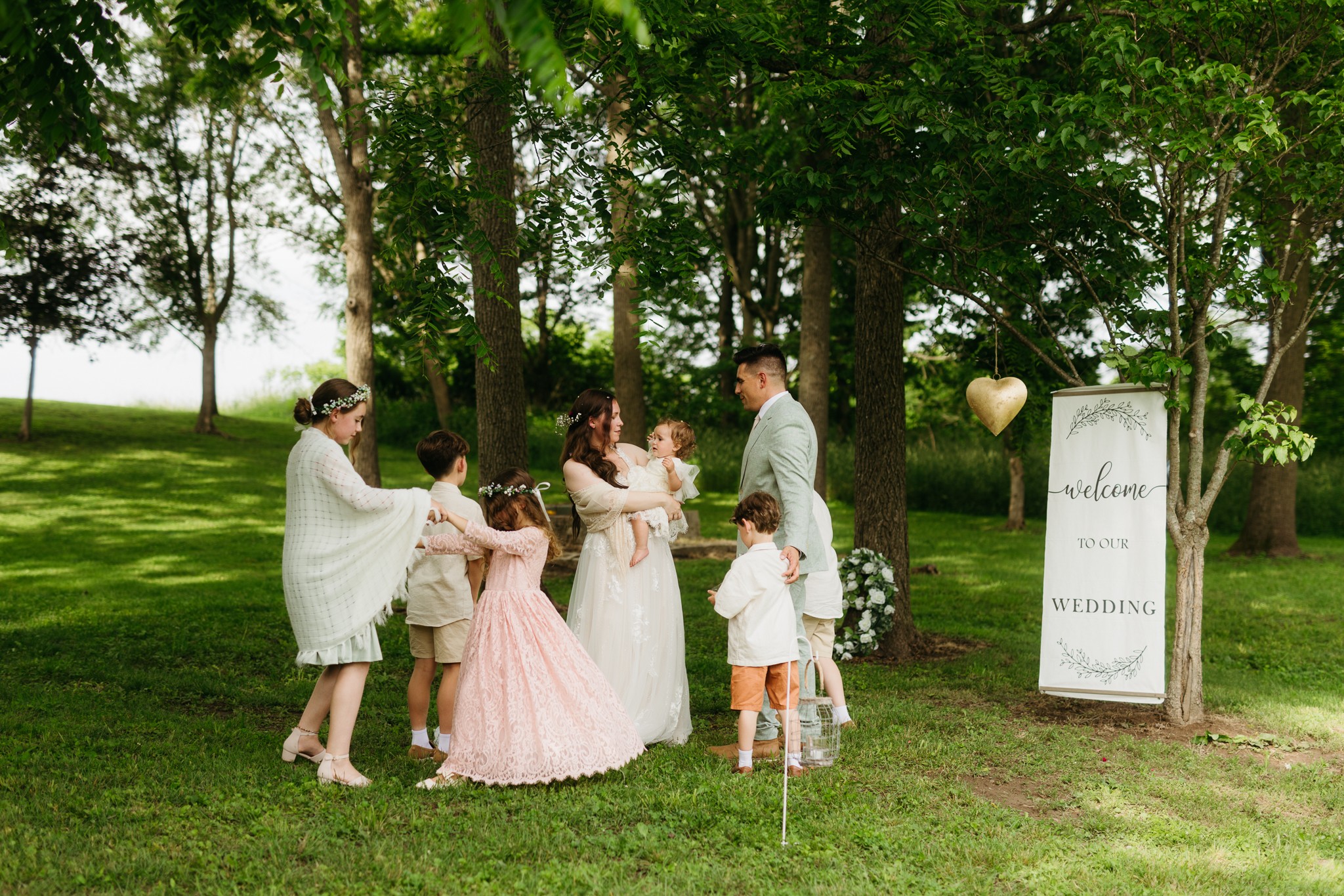 Group of people outdoors near trees, with a wedding sign and a heart-shaped balloon, celebrating a wedding.