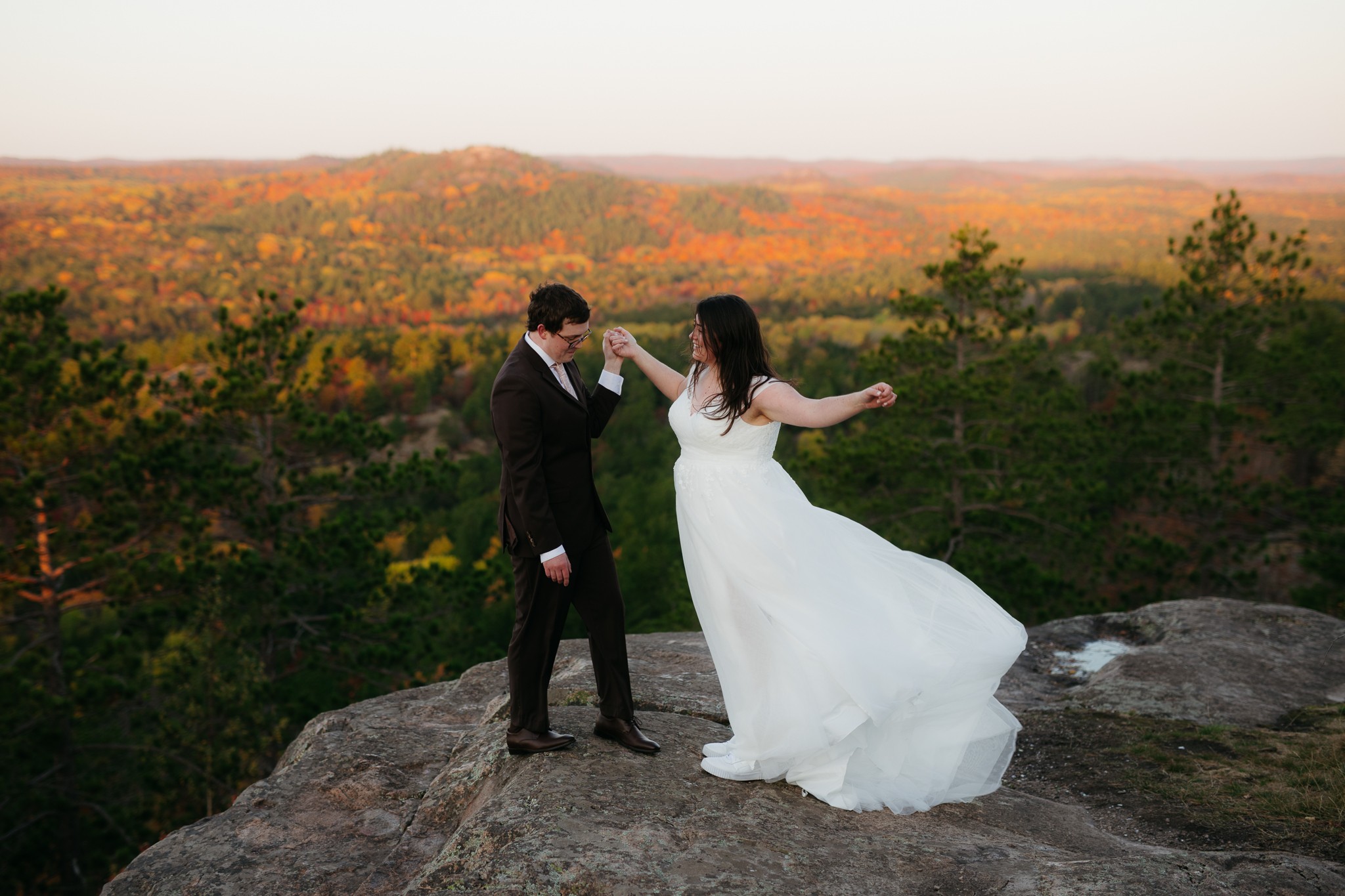 A couple in wedding attire standing on a rock with a scenic landscape of trees and hills in the background.