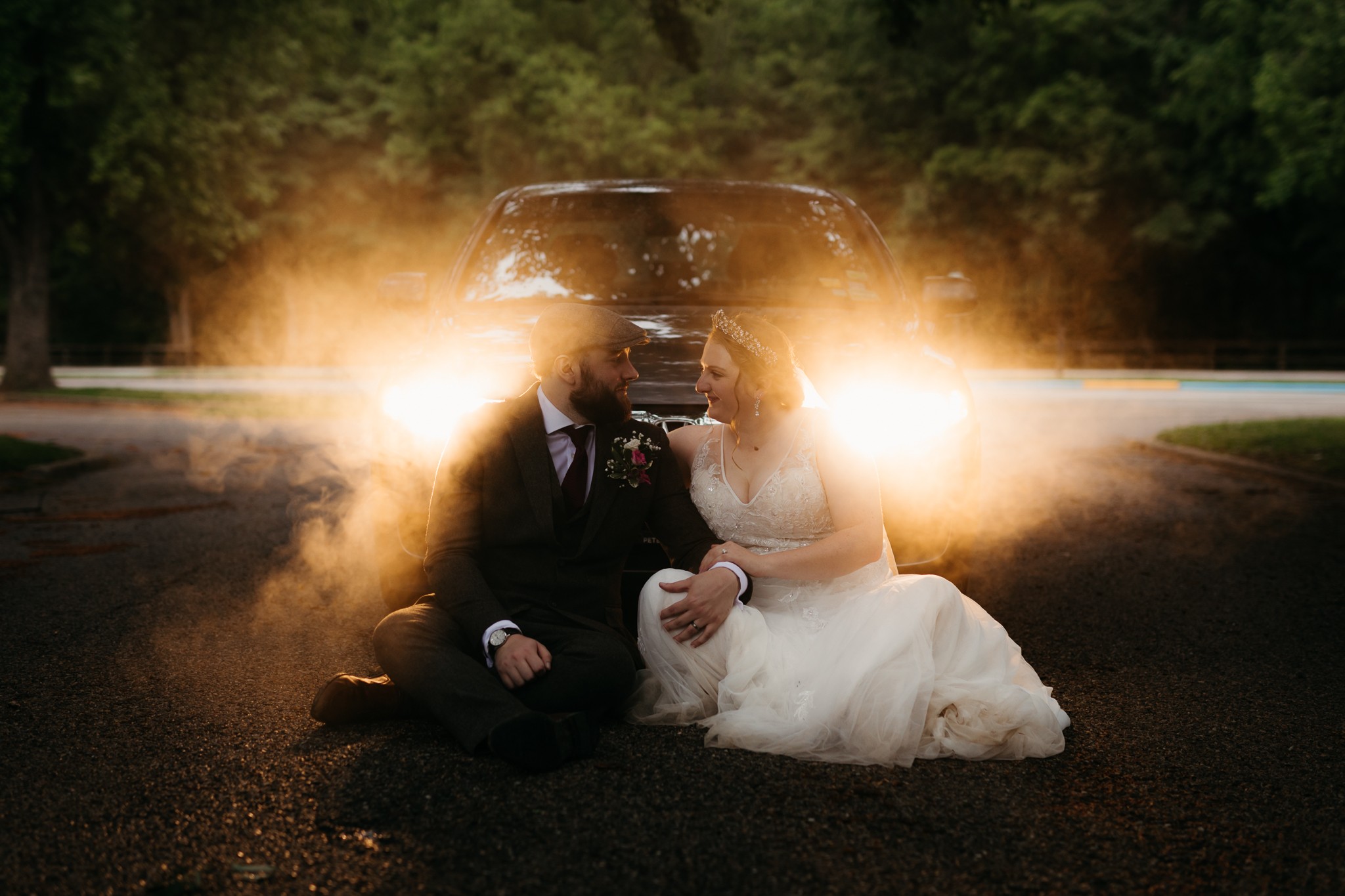 A bride and groom sit on the road with car headlights behind them, creating a bright glow and dust cloud. Wedding attire visible.