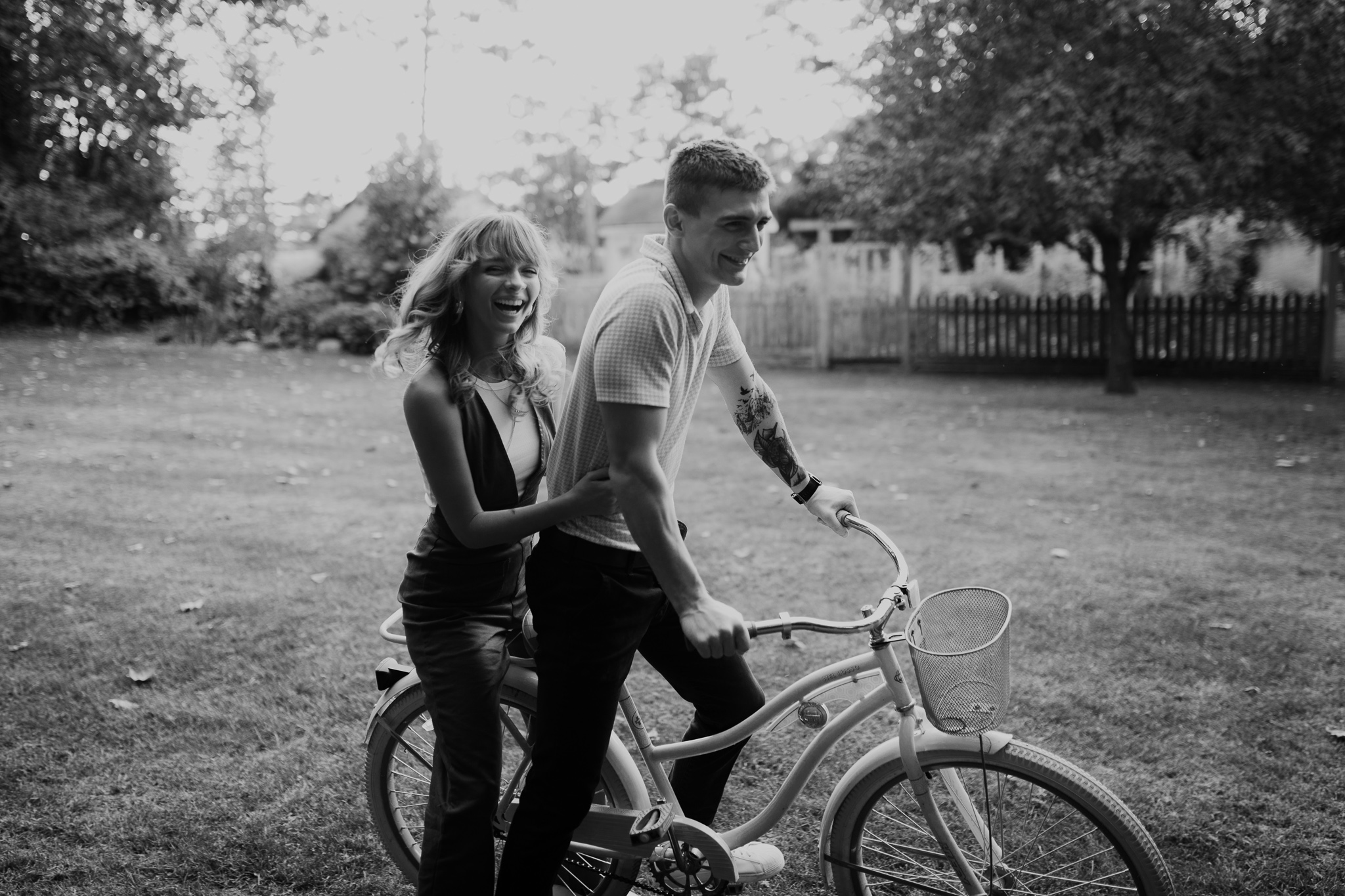Two people, a young man and woman, stand with a bicycle in a park with trees and a fence in the background.