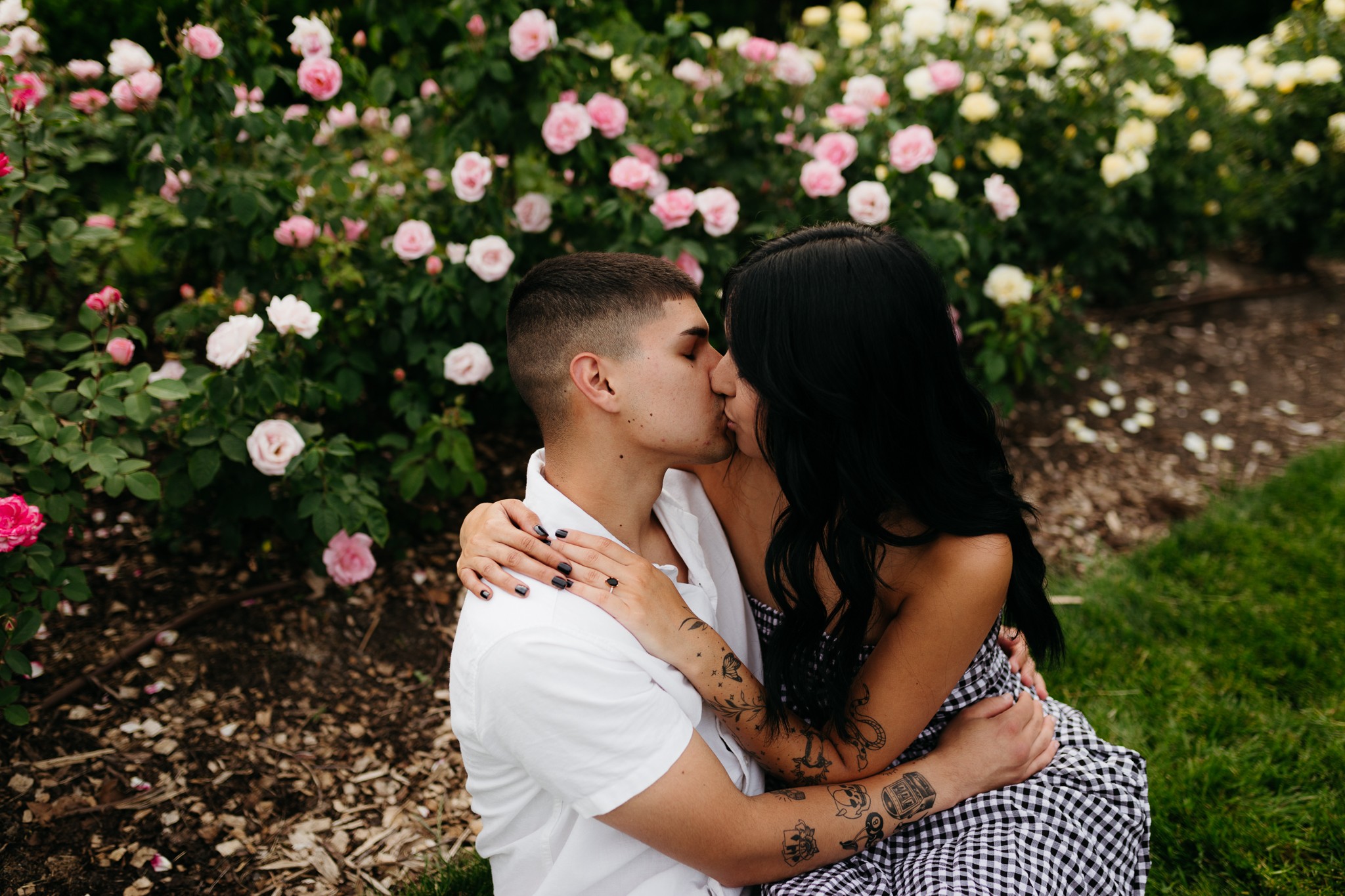 Two people kissing outdoors near pink and white flowers, one with short hair and the other with long dark hair, embracing.