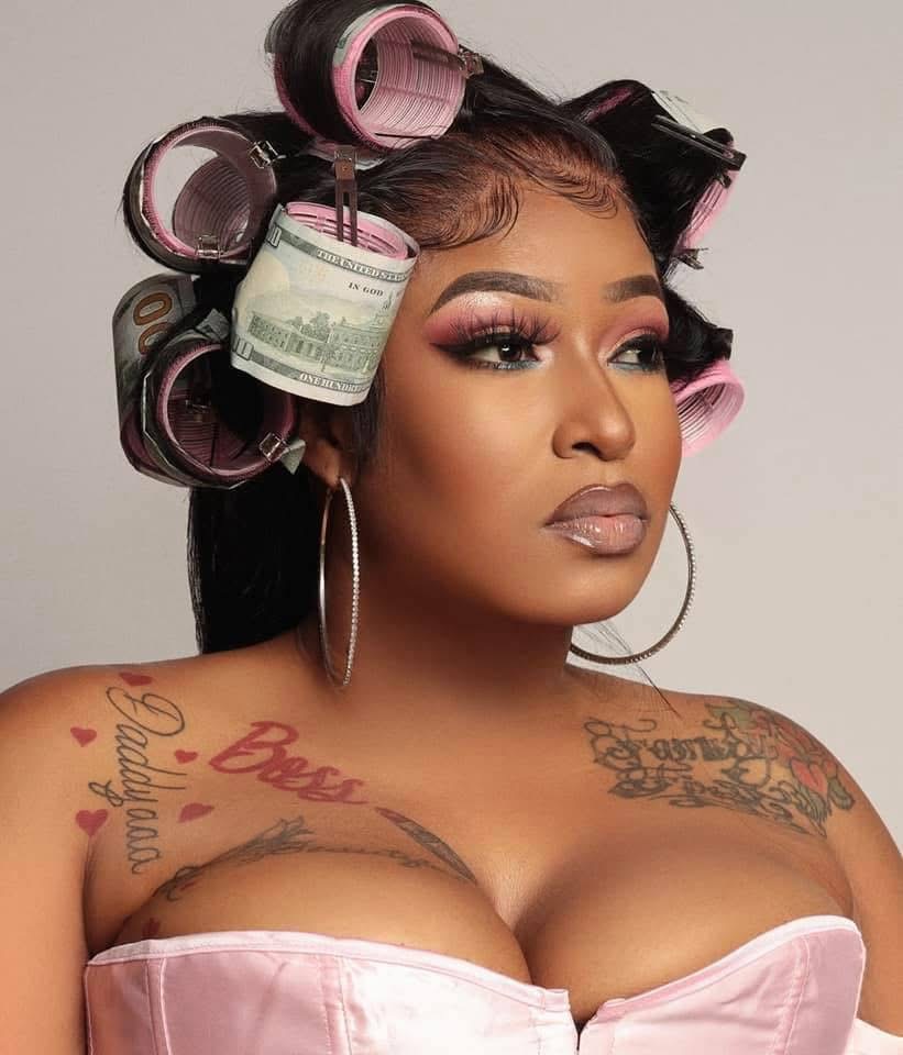 Woman with hair rollers, makeup, hoop earrings, and tattoos on shoulders and chest, wearing a pink strapless top.