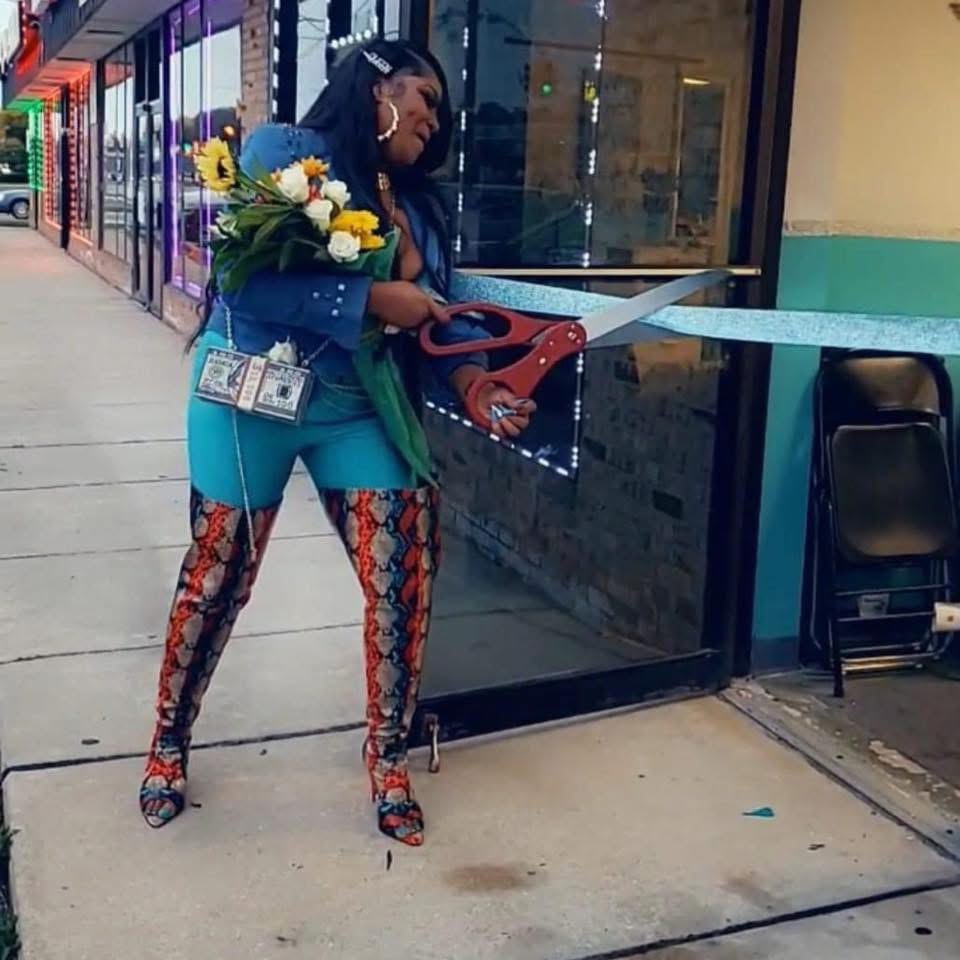 Woman with a bouquet of flowers and a large pair of scissors, standing outside a store, preparing to cut a ribbon.