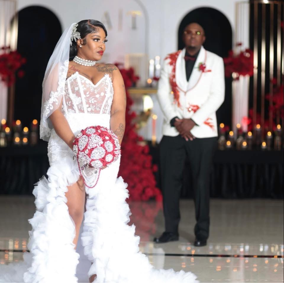 Bride in wedding dress holding bouquet, groom in white suit with red accents, wedding venue decorated with red flowers and candles.