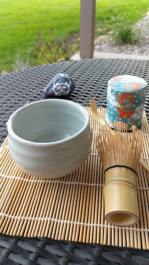 Ceramic bowl, bamboo whisk, and a decorated container on a bamboo mat outdoors, with grass and a post in background.