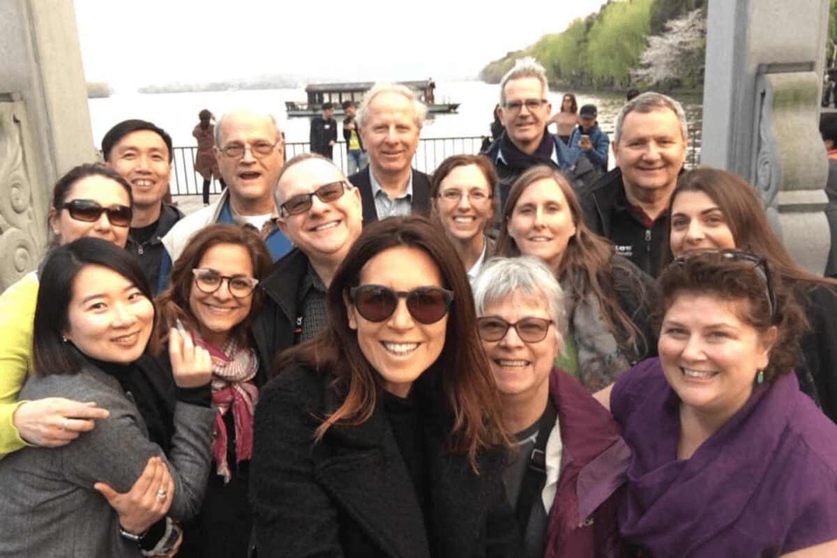 Group of 14 people smiling outdoors near a body of water and bridge, some wearing sunglasses, diverse ages and appearances.
