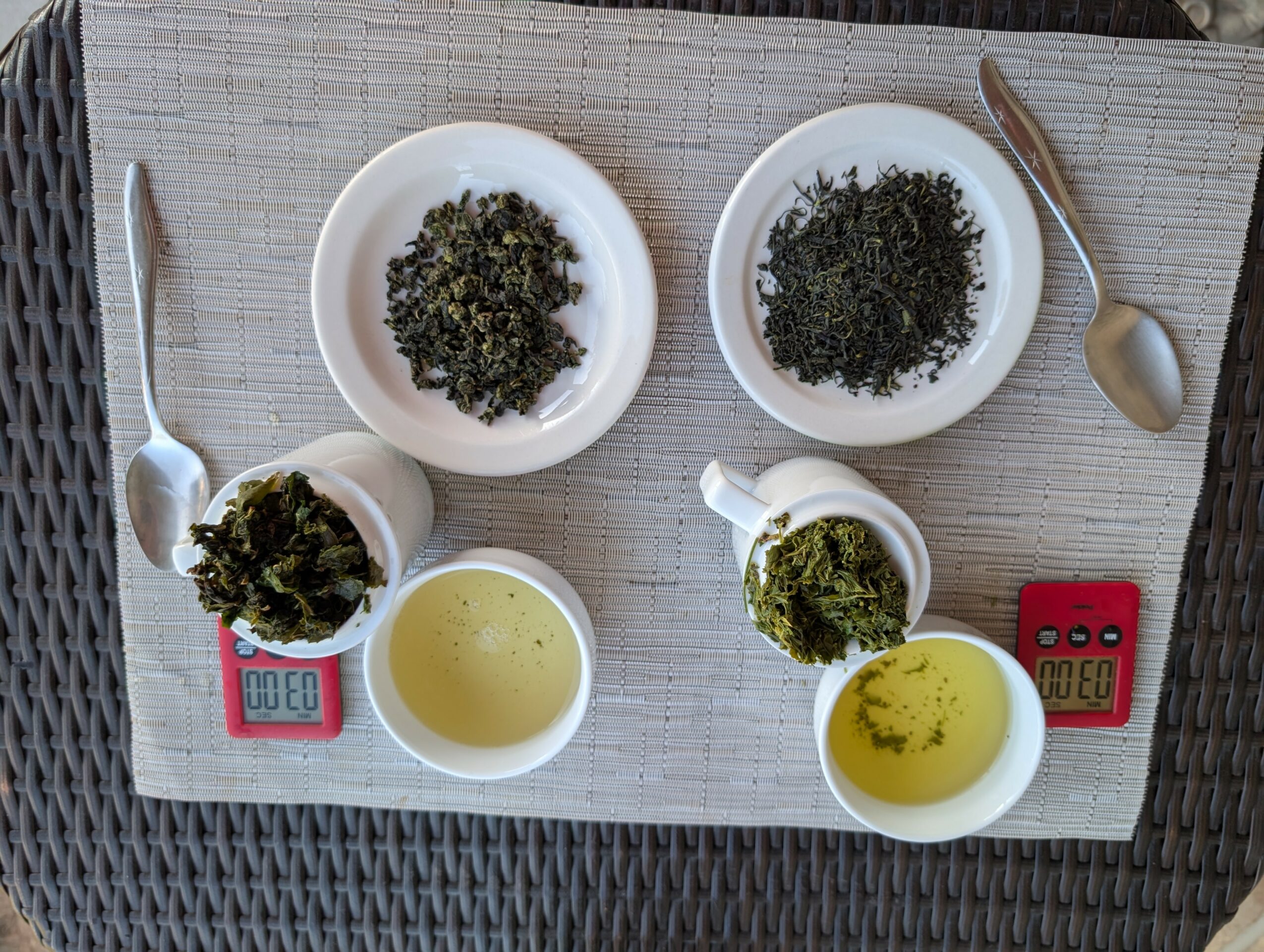 Two plates with dried herbs, two cups with liquid, and two spoons on a woven surface.