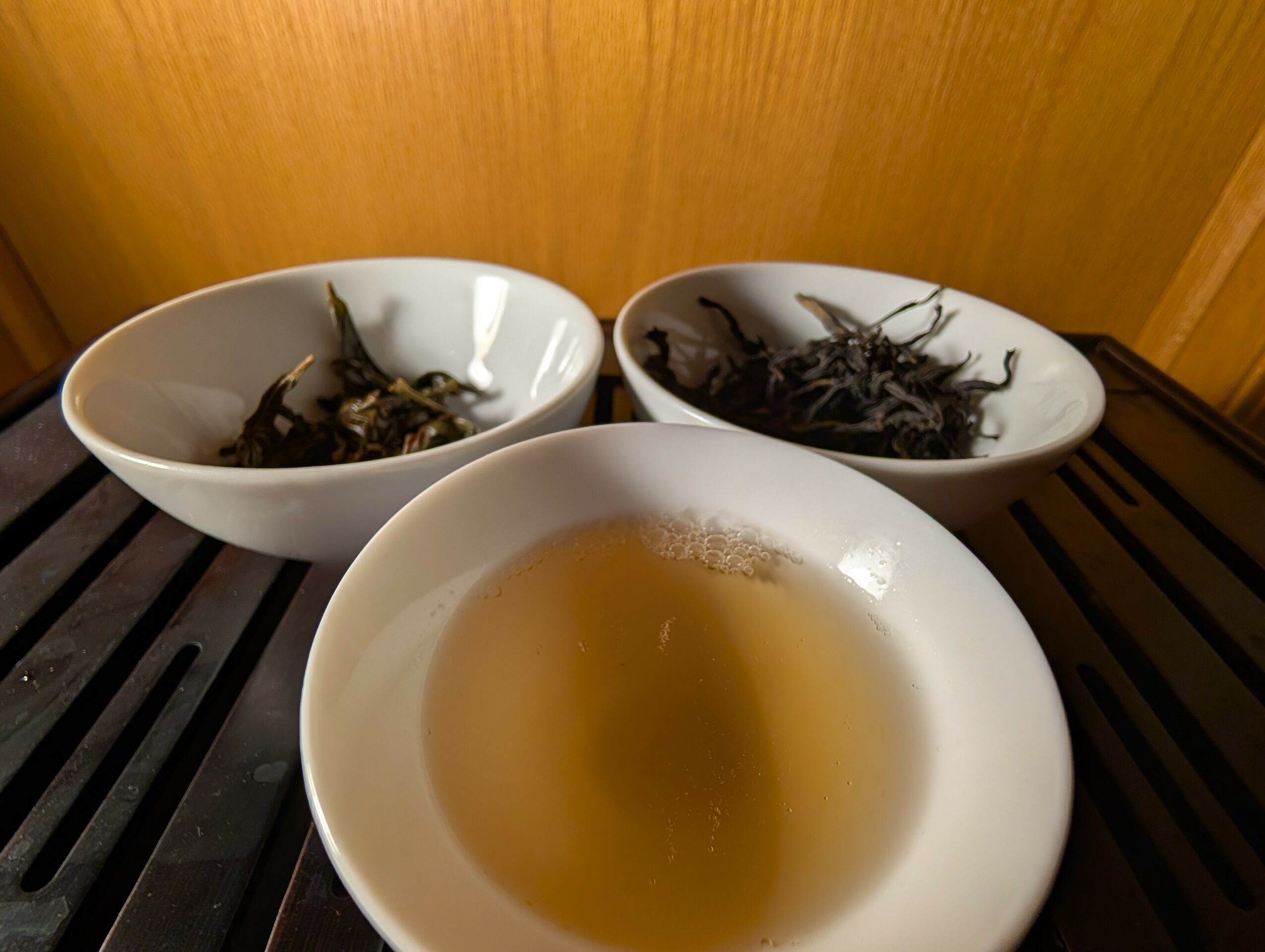 Three white bowls on a black surface, two with dried tea leaves and one with brewed tea, against a wooden background.