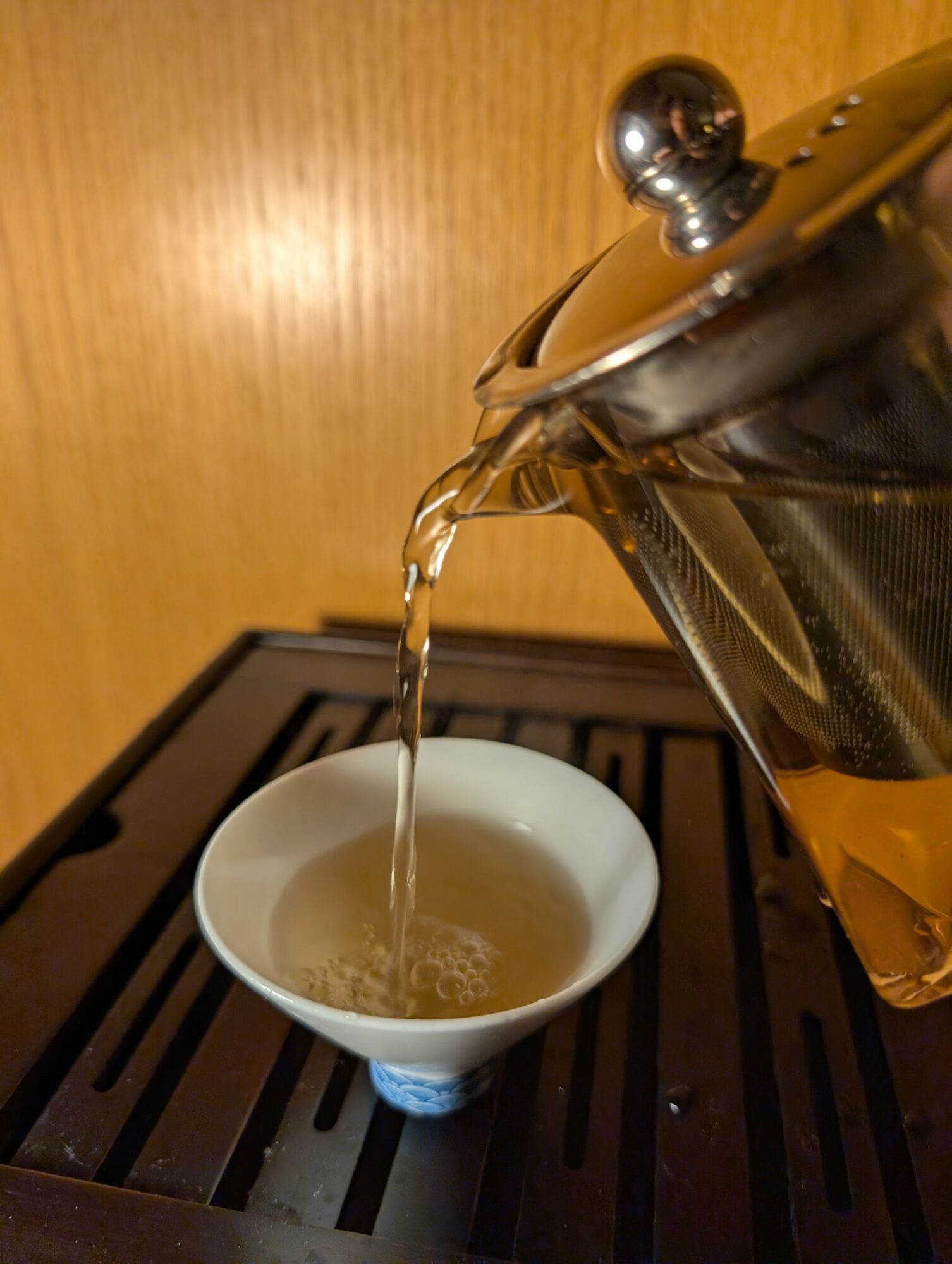 Pouring hot tea from a teapot into a small white cup on a wooden surface.