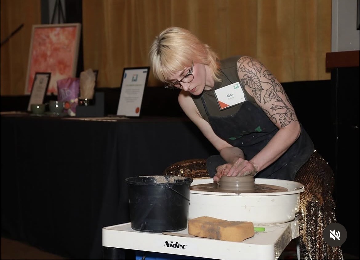 Woman with blonde hair and glasses shaping clay on a pottery wheel, wearing a black apron and lace sleeves.