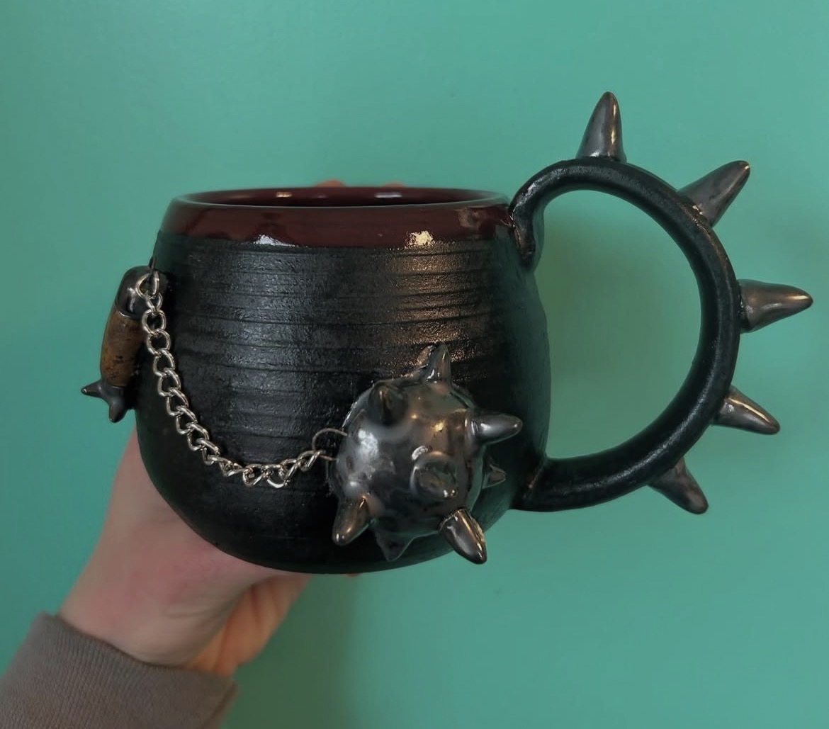 Hand holding a black mug with a spiked handle and a chain with a spiked ball, against a teal background.