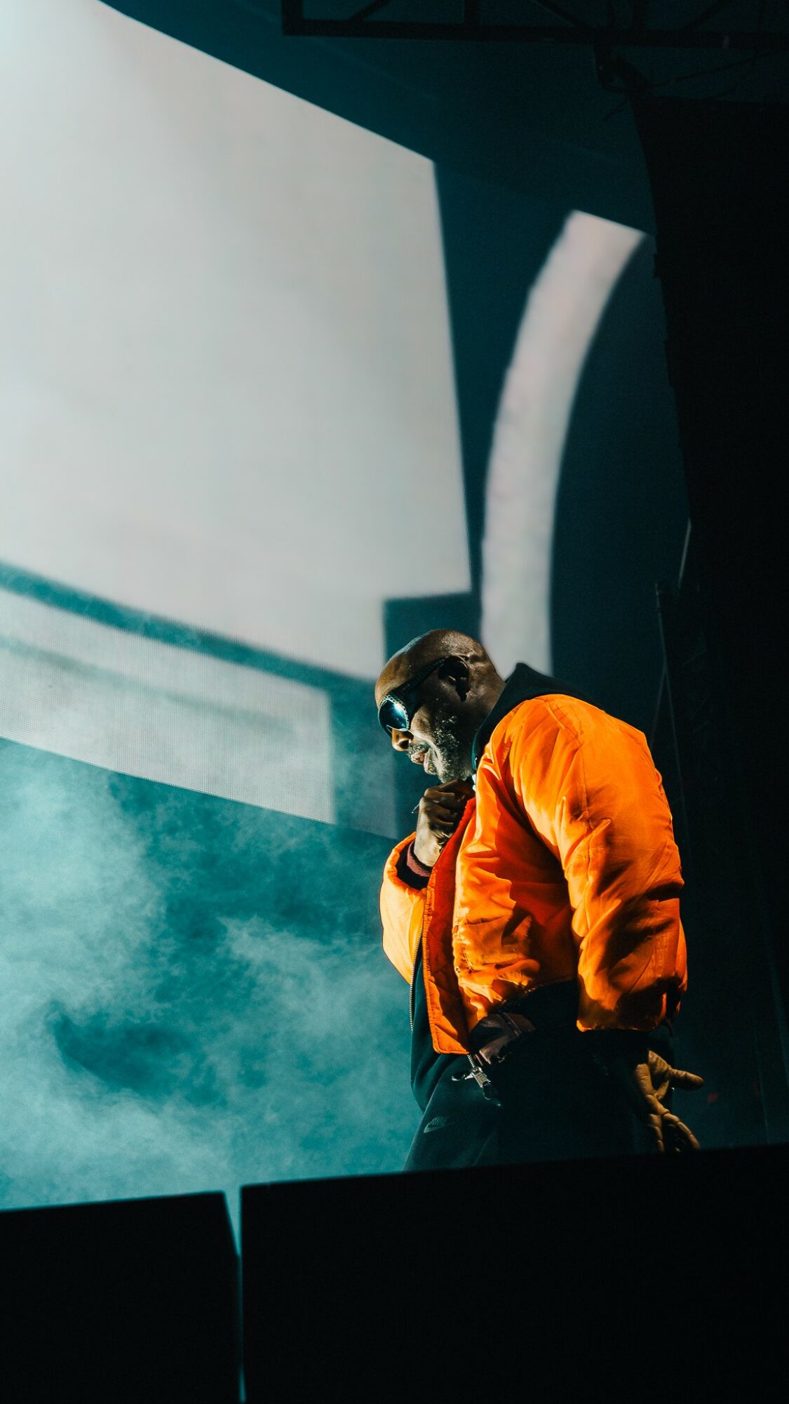 Person wearing orange jacket and sunglasses standing on stage with a large screen and smoke in background.