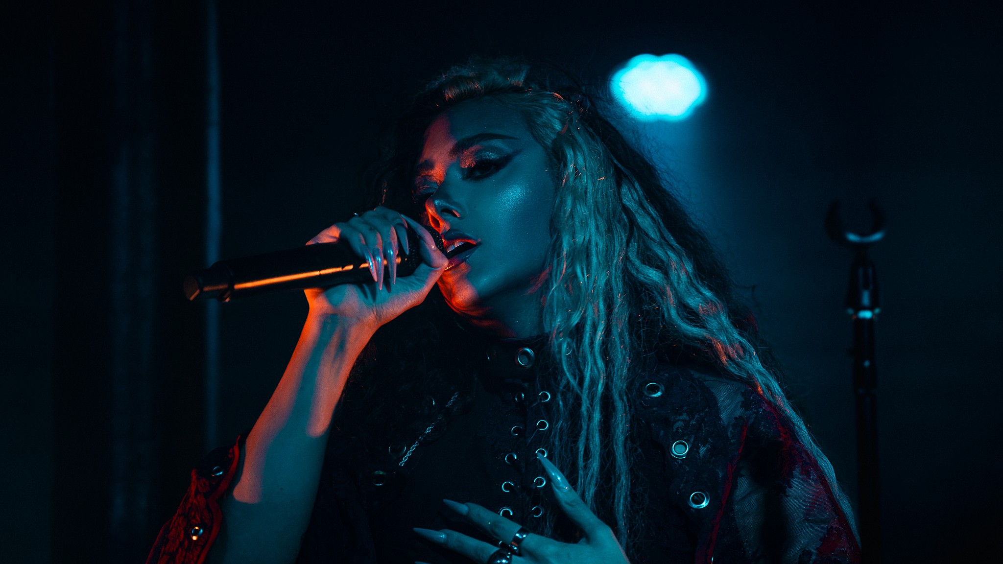 A woman singing into a microphone on stage with blue lighting, holding her chest with one hand.