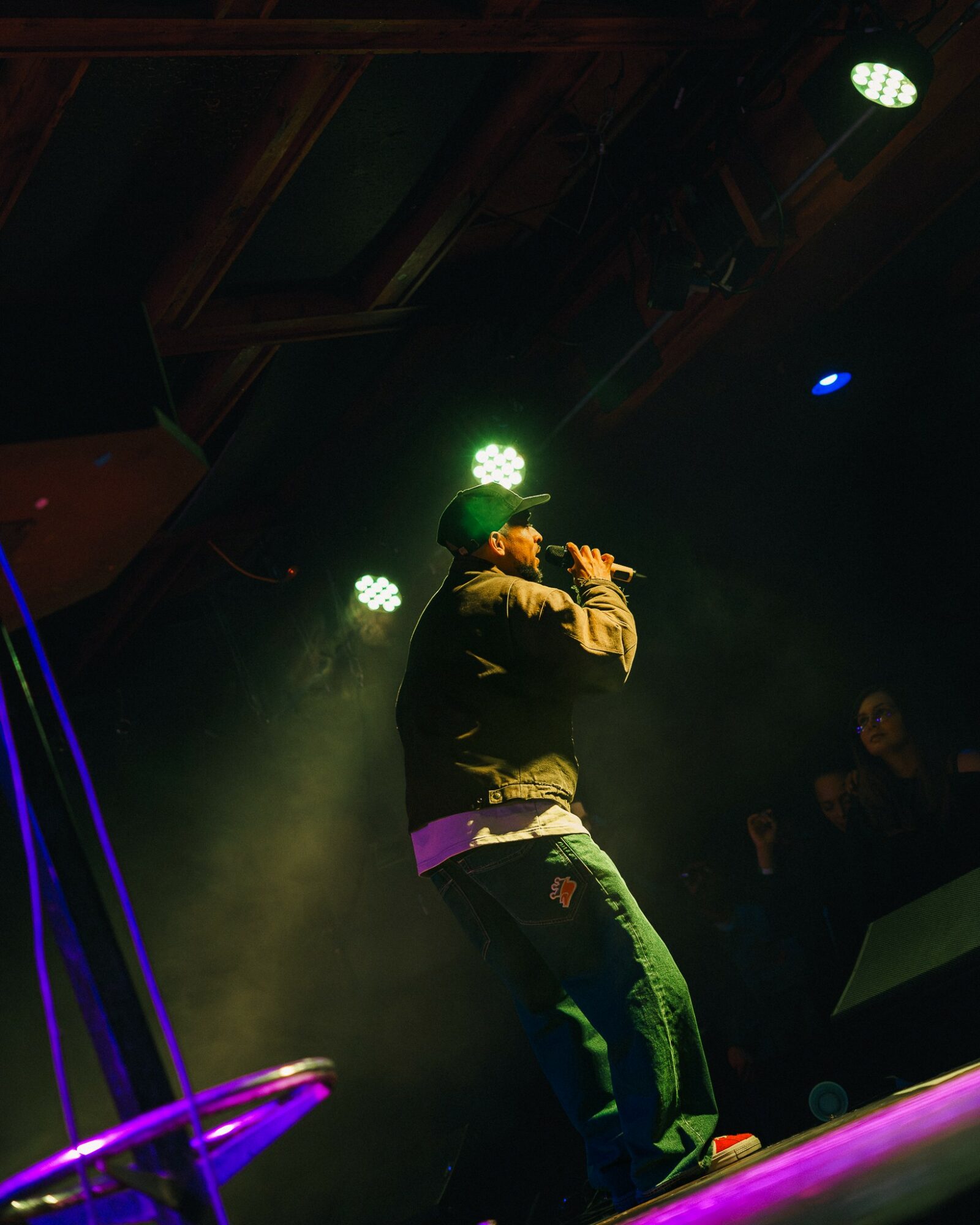 Performer on stage holding microphone, wearing a hat and casual clothing, with stage lights and audience in background.