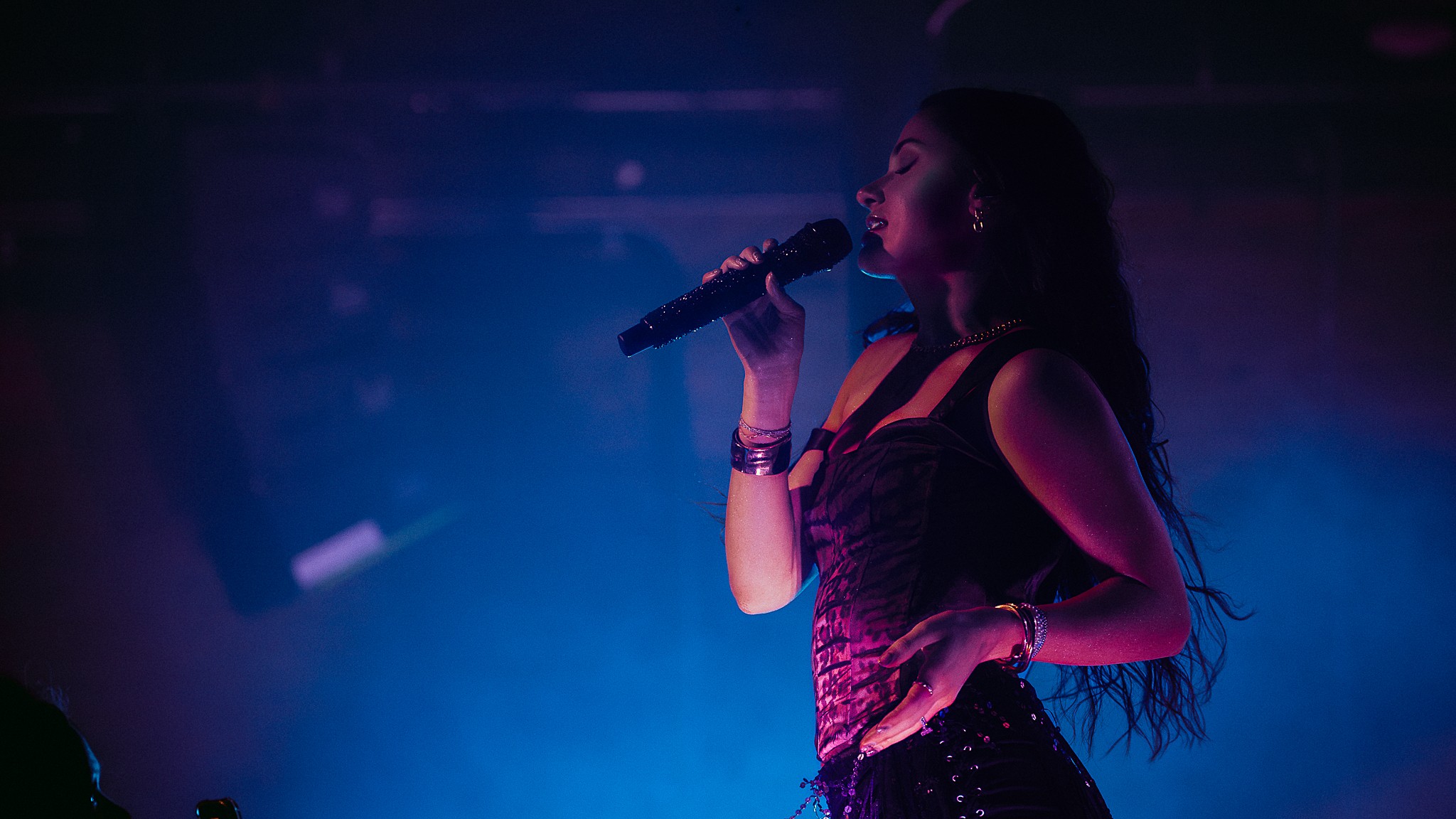A woman singing into a microphone on stage with blue and purple lighting, facing right side, with long hair and a sleeveless top.