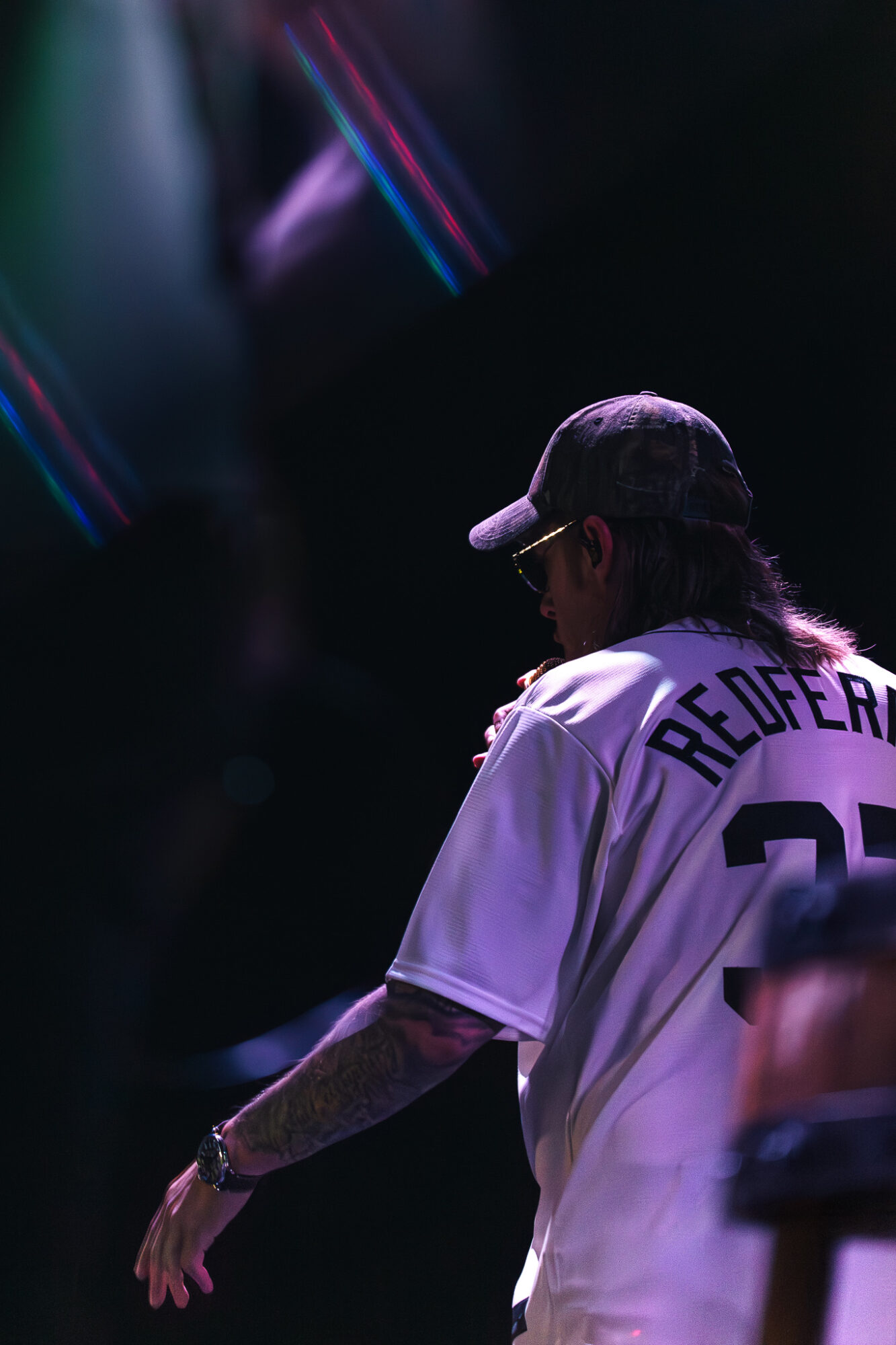 Person wearing a baseball jersey and cap, facing sideways, with dark background and colorful lights.