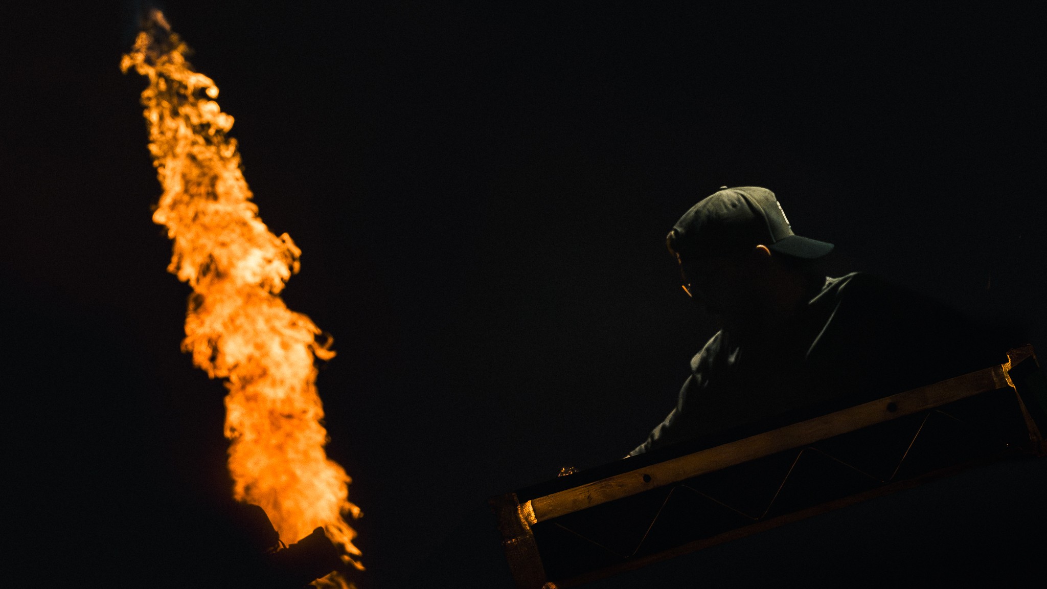 Person wearing a cap working near a flame or fire against a dark background.