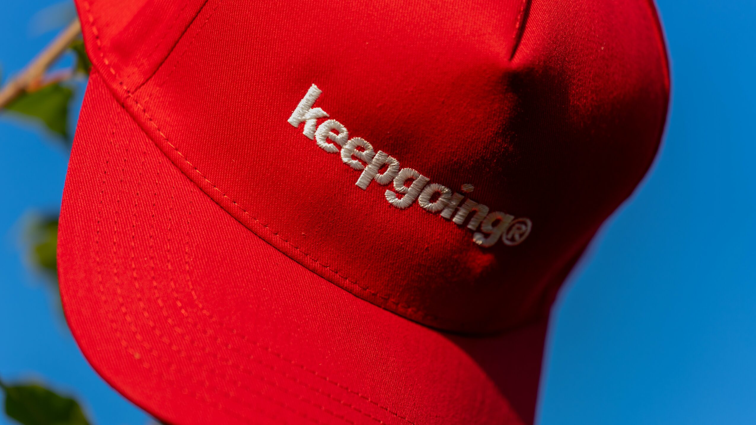 Red cap with white embroidered text 'keepgaming' against a blue sky background.