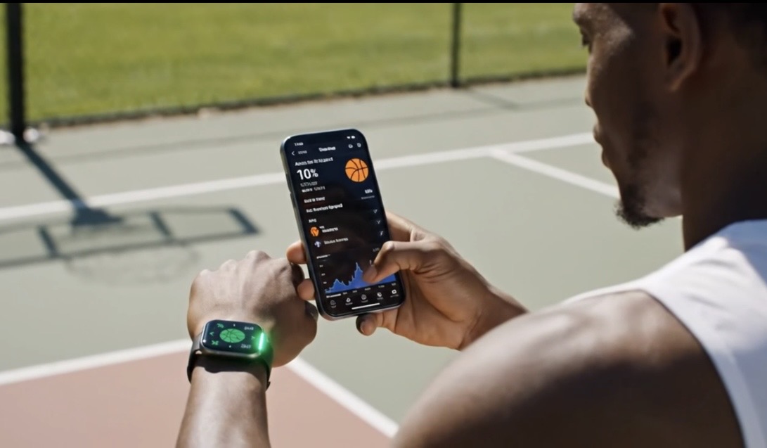 Person holding a smartphone outdoors on a tennis court, wearing a smartwatch, with a grassy area and fence in background.