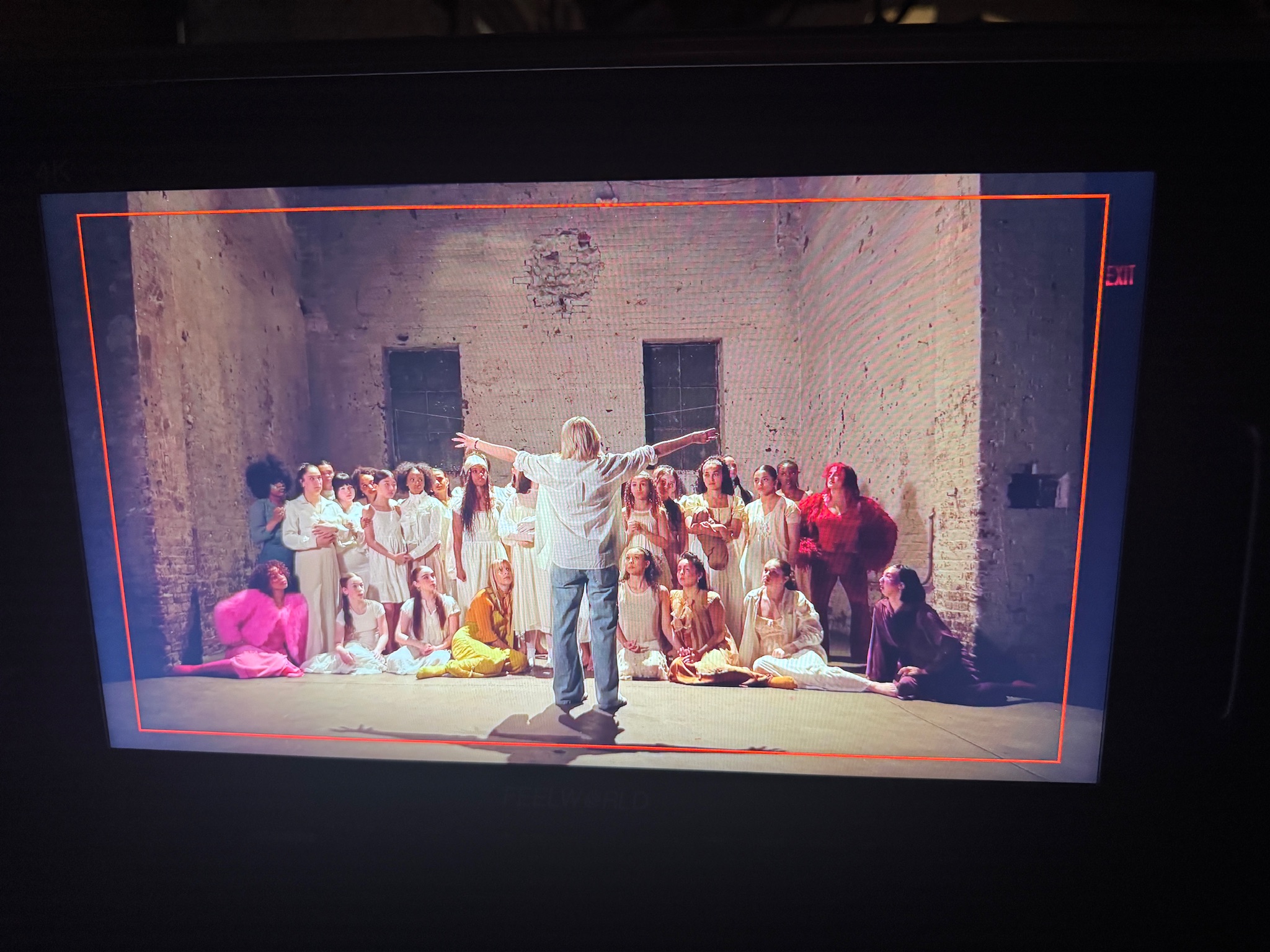 A group of children and adults on stage in a large, plain room with stone walls, with a person directing them.