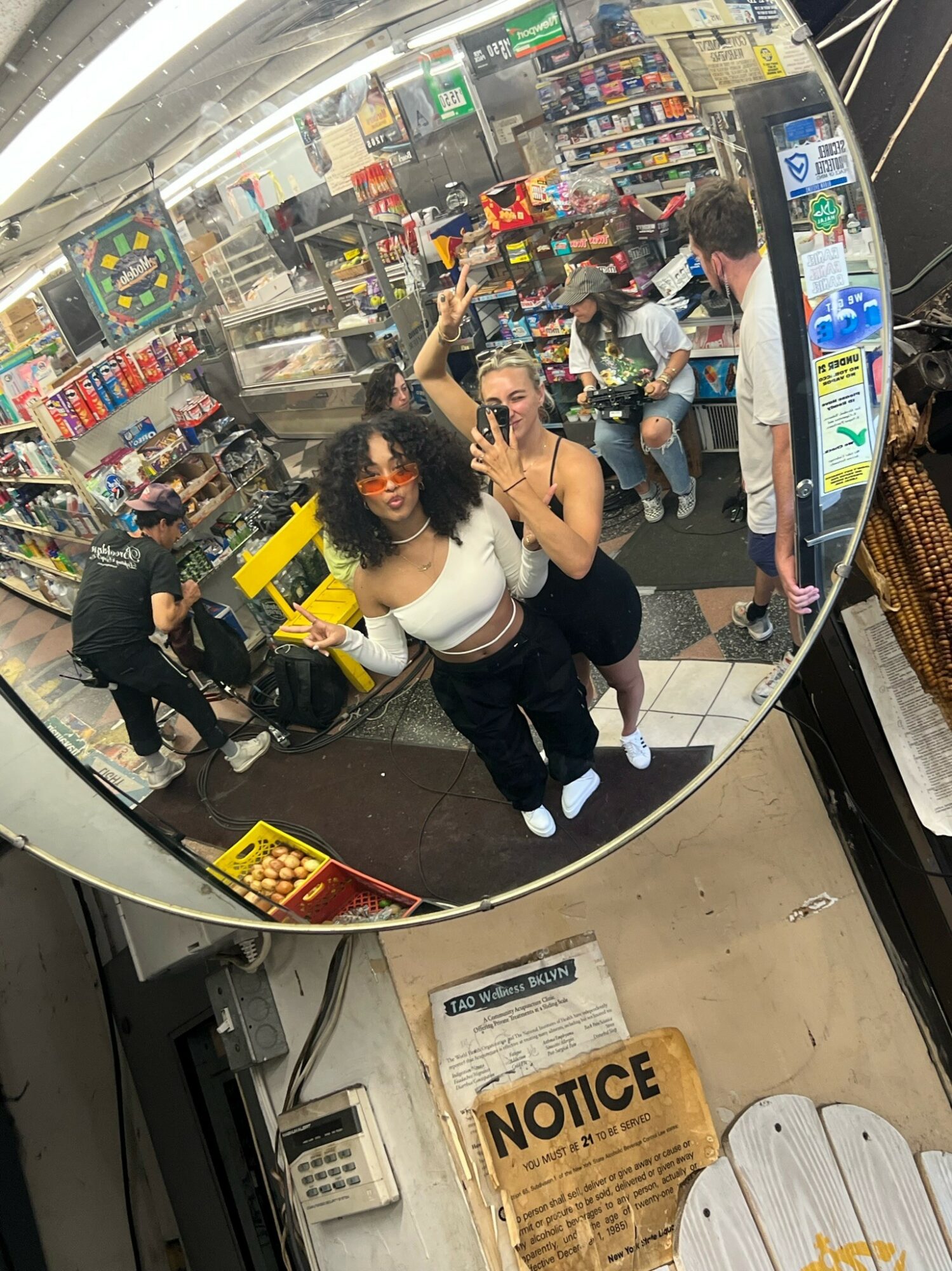 Two women taking a selfie in a store's mirror, surrounded by shelves of products and other shoppers.