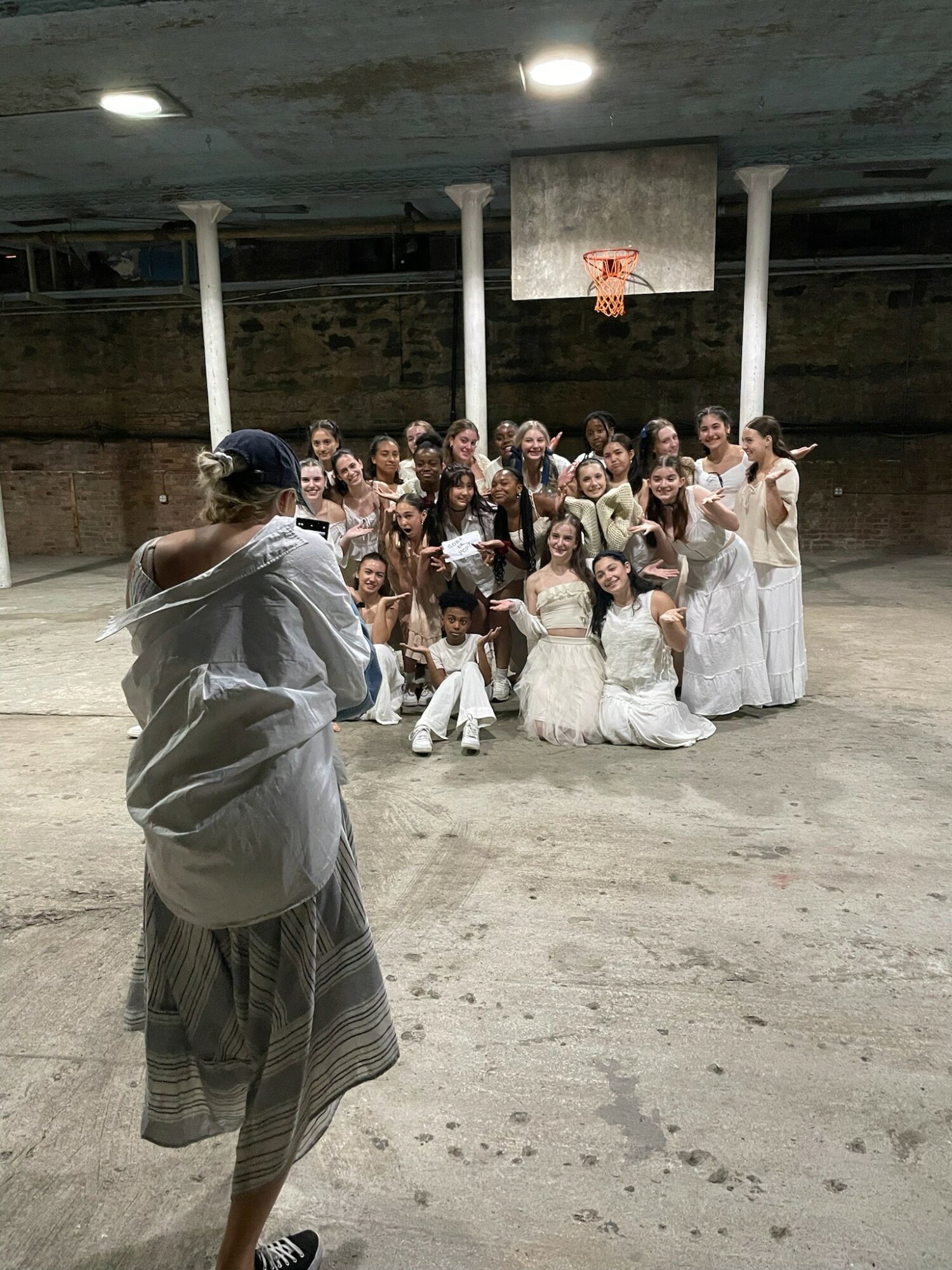 Group of people in white dresses posing for a photo in an indoor basketball court, with a person taking the picture in the foreground.