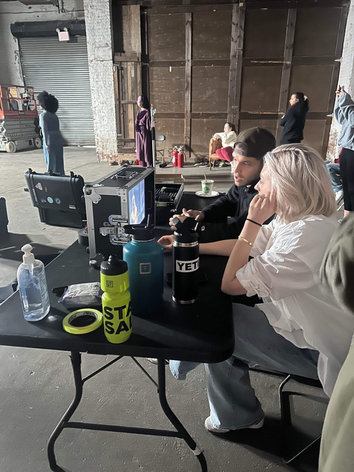 Two people sit at a table with computers and water bottles, while others stand in the background in an industrial setting.