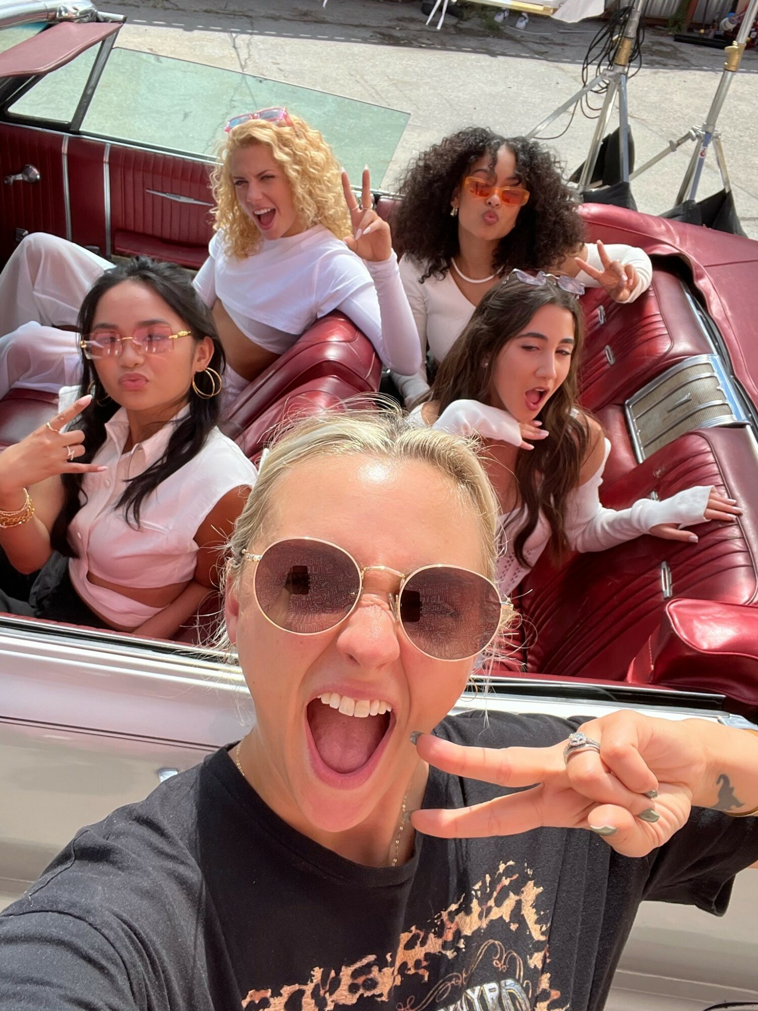 Five women in a convertible car, smiling and making peace signs, enjoying a sunny day.