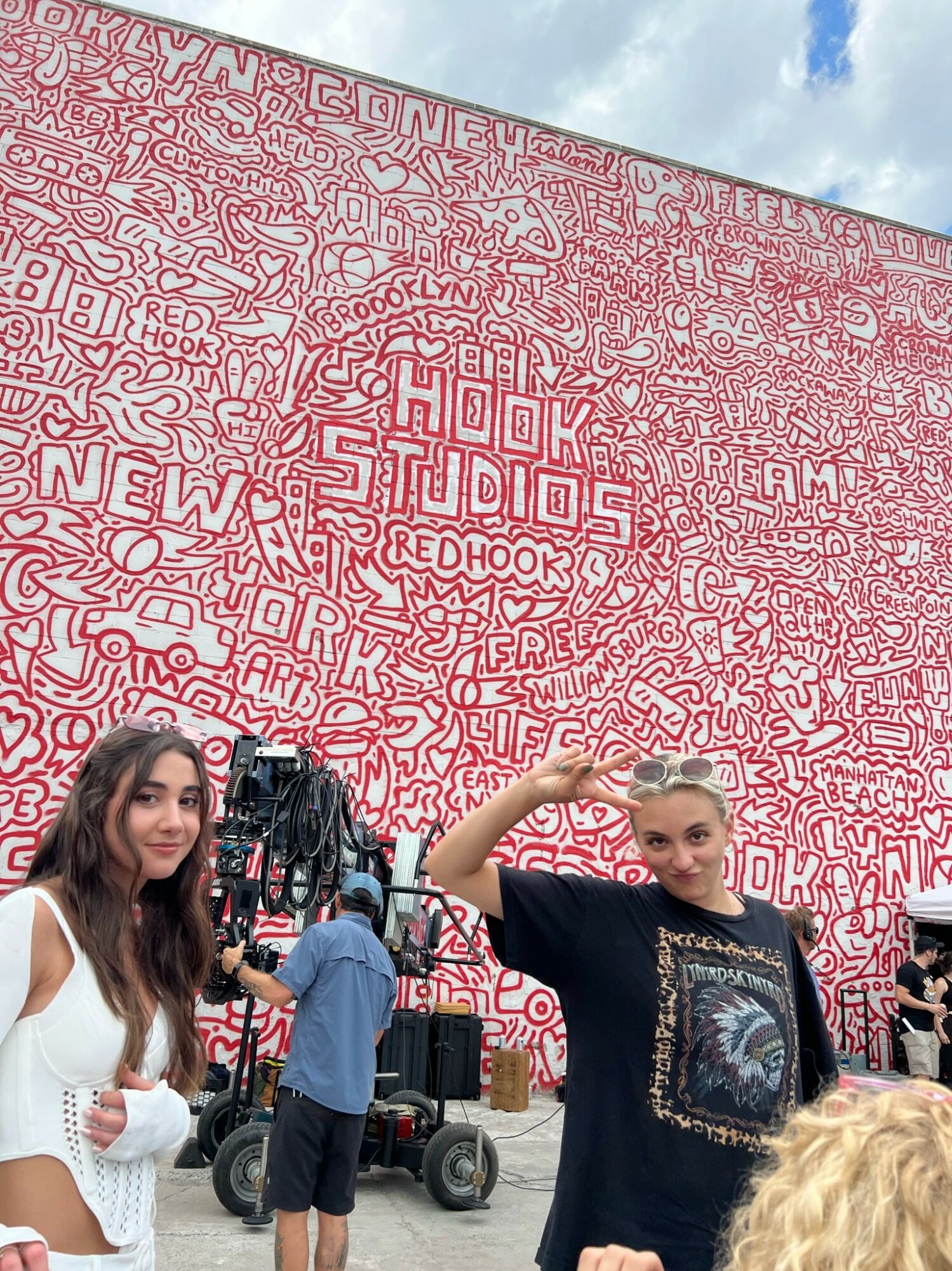 Two women standing in front of a large pink wall with white text and patterns, one saluting, the other holding a bag.