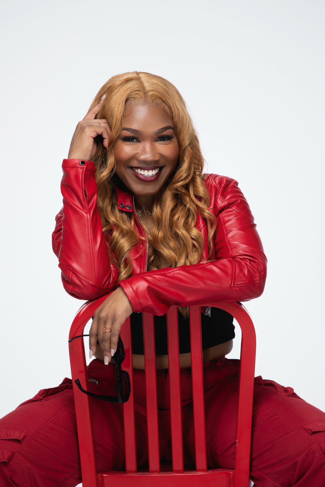 Smiling woman with long blonde hair in a red jacket and pants, sitting on a red chair, against a white background.