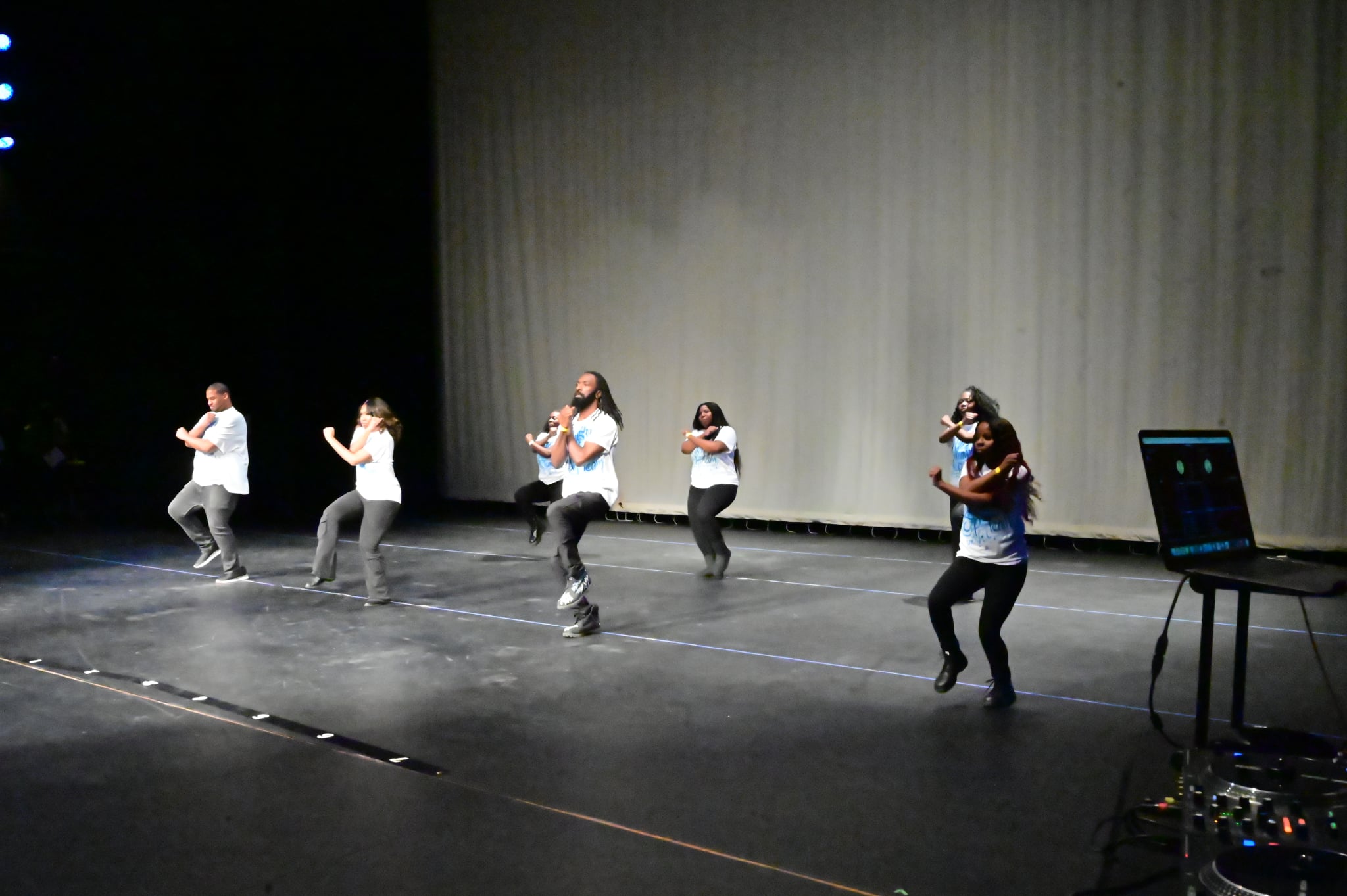 Six people dance on stage with a plain curtain background, some wearing masks, with a laptop on a stand nearby.