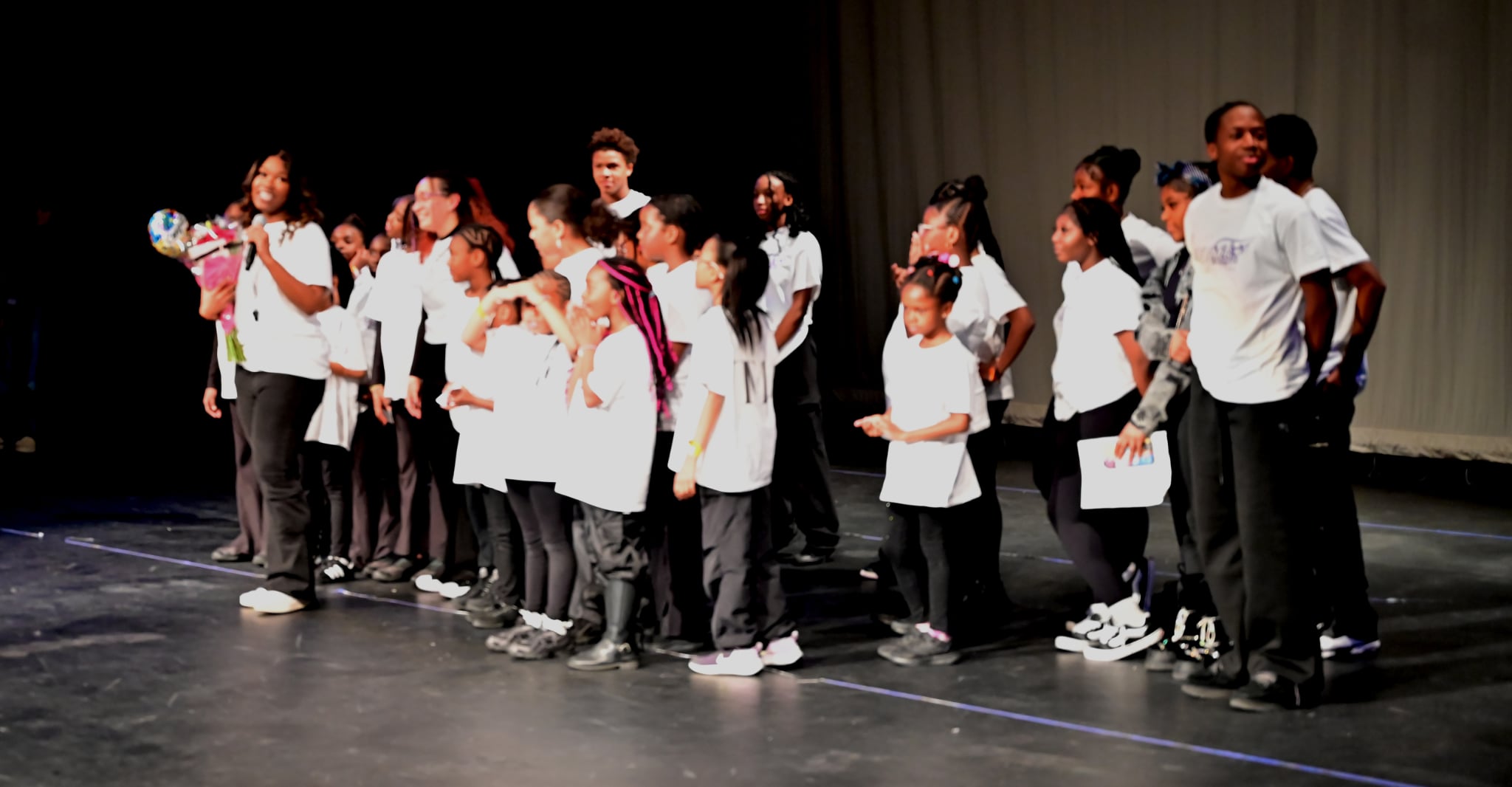 Group of children standing on stage, some holding flowers, wearing white shirts and dark pants, in front of a curtain.