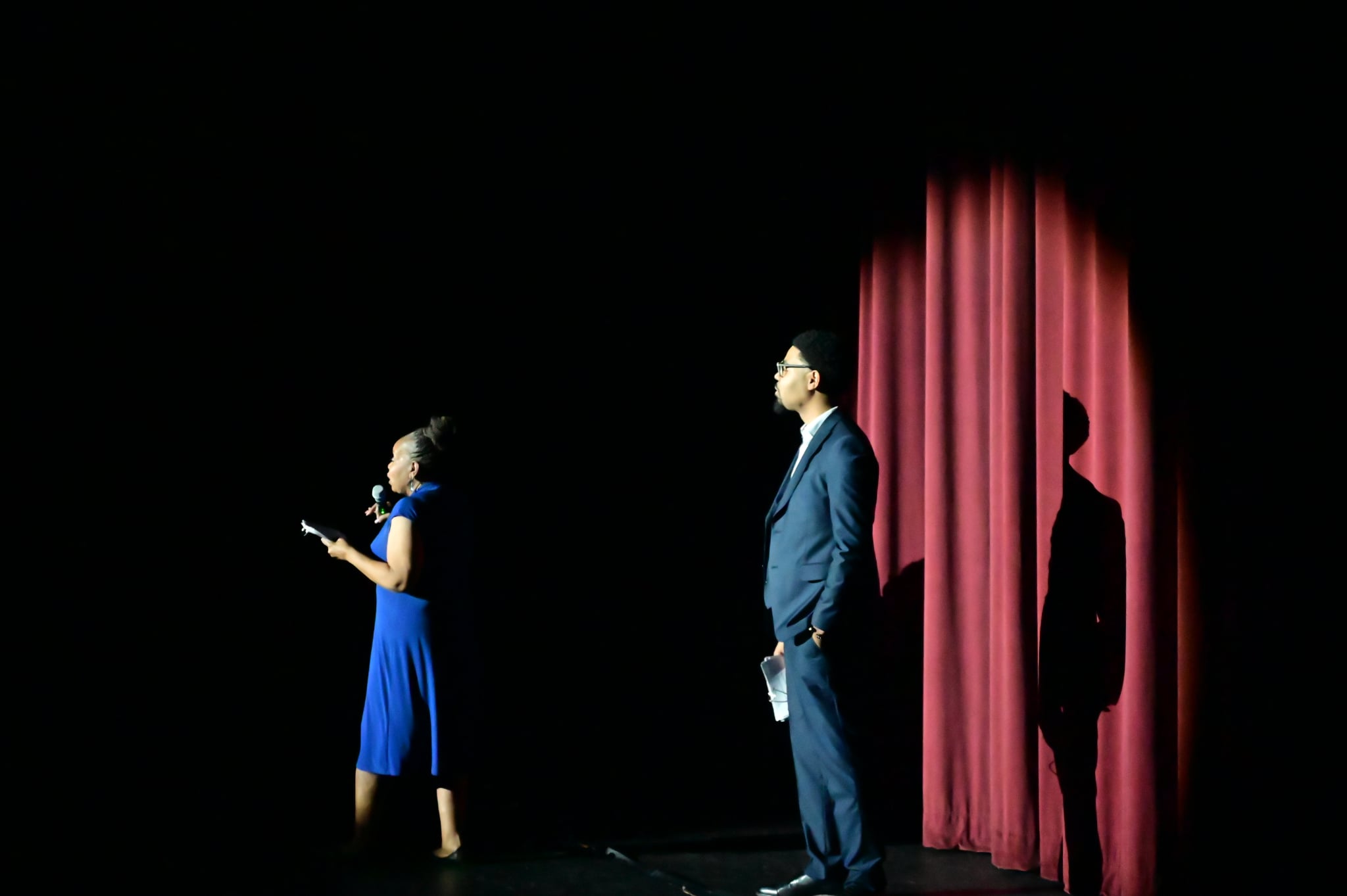 Person in blue dress reading on stage, man in suit waiting behind red curtain, shadow cast on curtain, dark background.