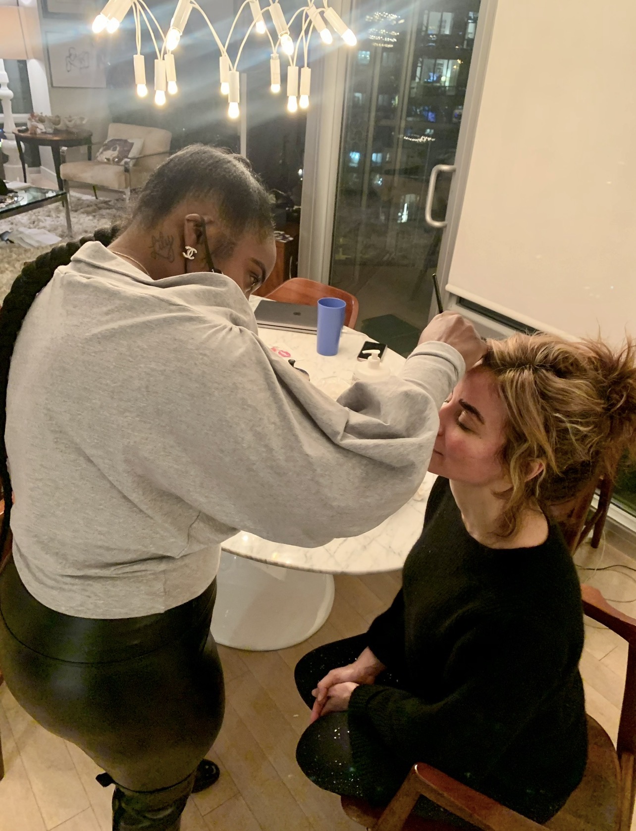 Makeup artist applying makeup to a seated woman in a room with large windows and hanging lights.