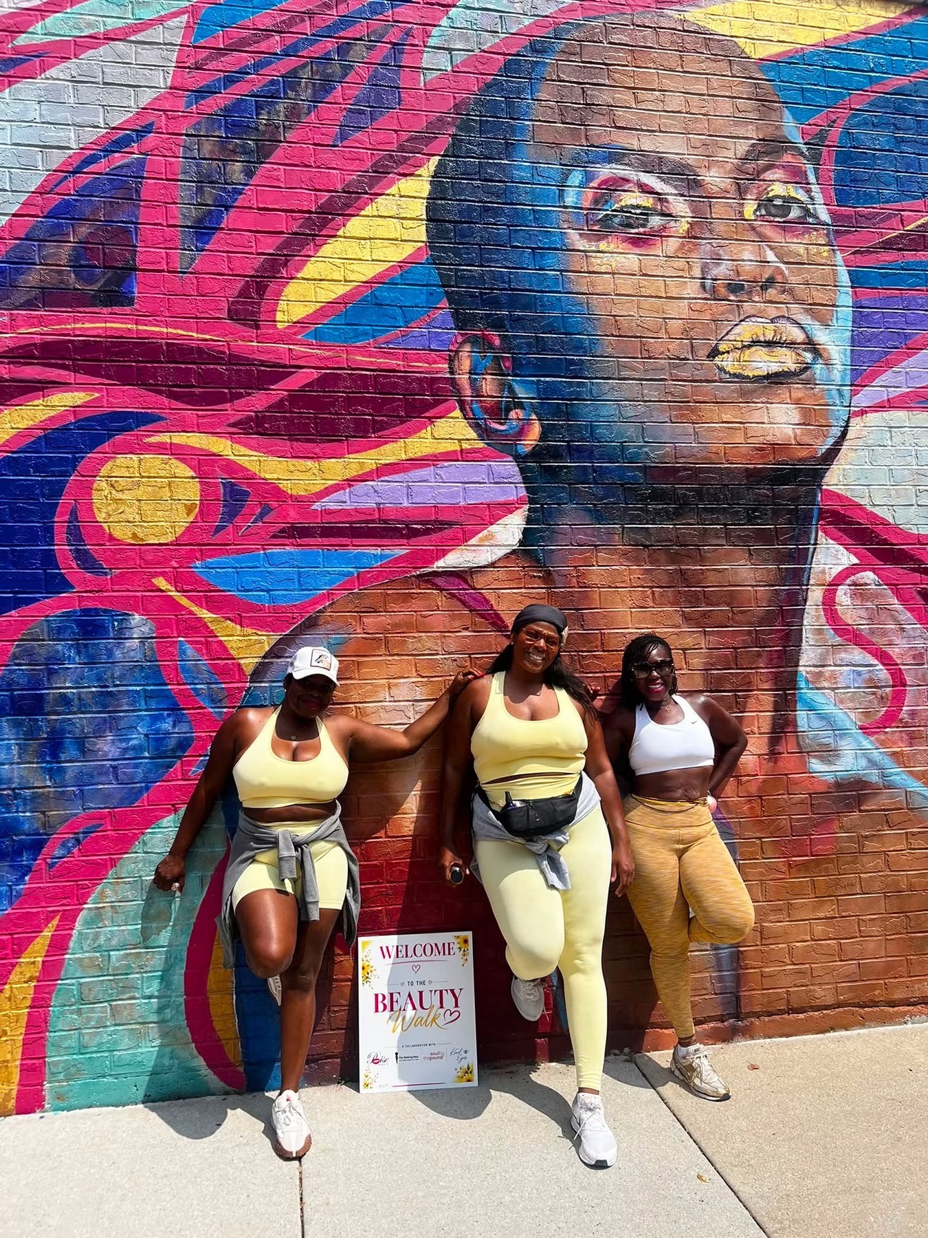 Three women stand in front of a colorful mural of a woman's face with vibrant hair, posing for a photo.