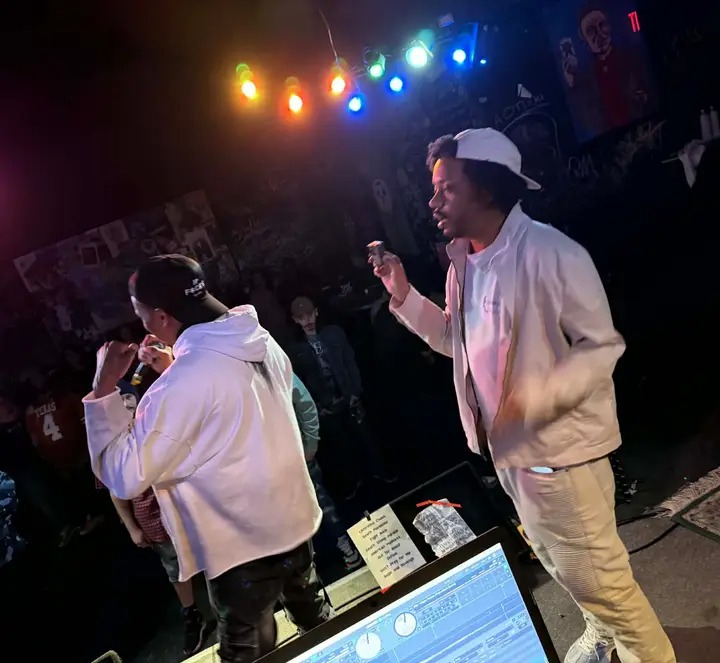 Two people in white hoodies and caps are on stage with colorful lights overhead, engaging with the audience.