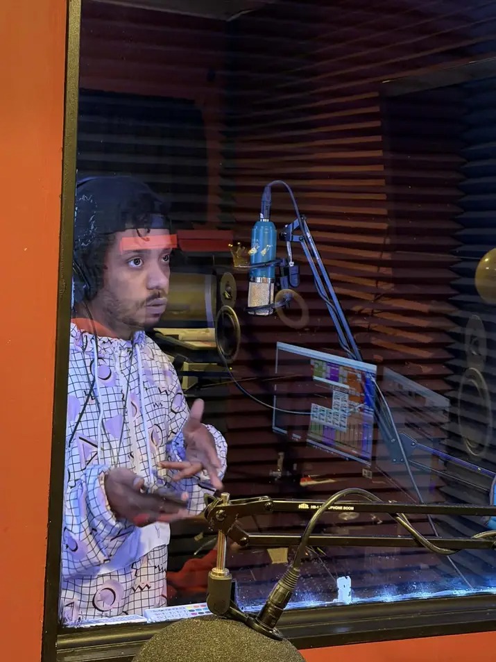 Person with curly hair in a recording studio, wearing a patterned shirt, looking through a glass window, with audio equipment visible.