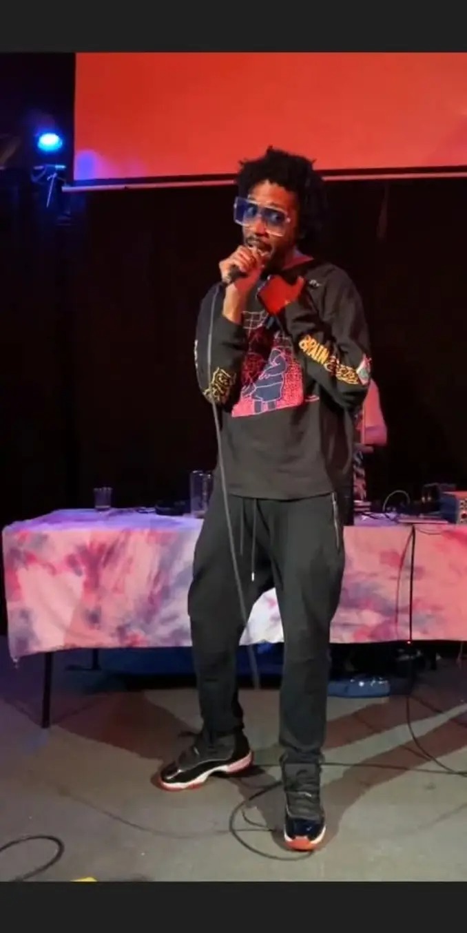 Person with curly hair and sunglasses singing into a microphone on stage, with a red background and a table with a pink cloth.